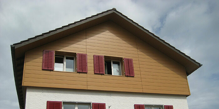 Rivestimento del timpano di una casa tradizionale con tetto a due falde. Il timpano è rivestito con doghe di rivestimento PREFA (rovere naturale) in posa orizzontale. Le finestre sono provviste di persiane rosse.