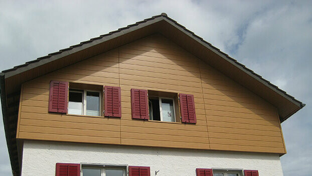 Giebelverkleidung eines klassischen Hauses mit Satteldach. Der Giebel ist mit horizontal verlegten PREFA Sidings in Eiche natur verkleidet. Die Fenster haben rote Fensterläden.
