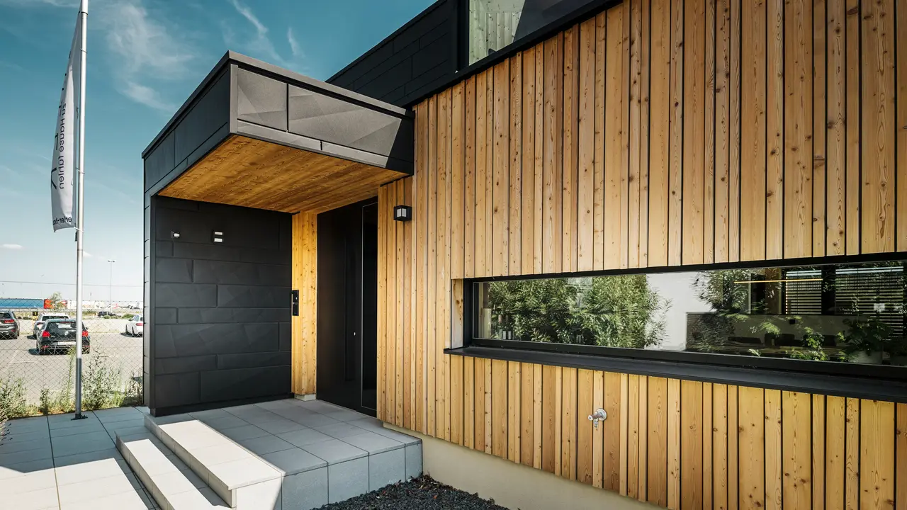Ingresso laterale della casa campione Autark nel centro prefabbricati Blaue Lagune a Vösendorf, con una facciata moderna in legno naturale e  Doghe.X PREFA. L'immagine mostra la raffinata combinazione di rivestimento in legno e alluminio nel pregiato rivestimento P.10 nero.