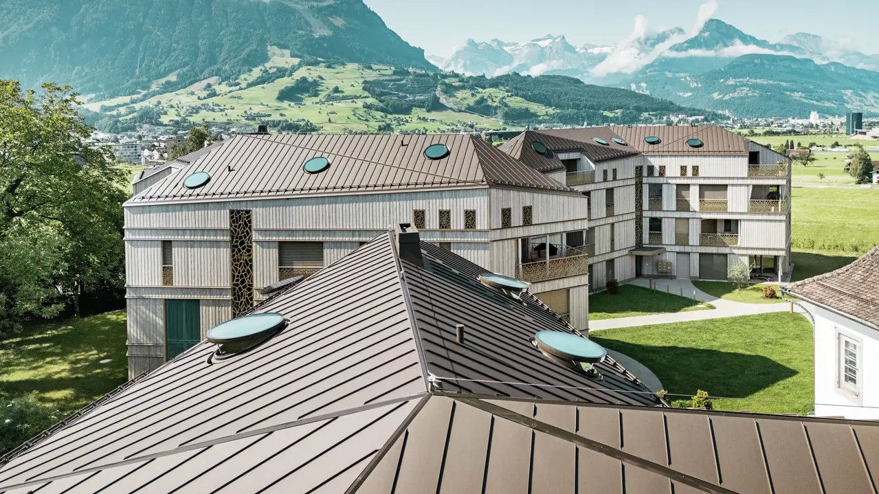 Moderno complesso residenziale a Liebwylen, in Svizzera, con un innovativo paesaggio di tetti in alluminio PREFALZ di colore marrone.  Il tetto con tecnologia a doppia aggraffatura integra numerosi lucernari rotondi e si inserisce armoniosamente nel paesaggio montano circostante. Sullo sfondo, un imponente paesaggio alpino e prati verdi.