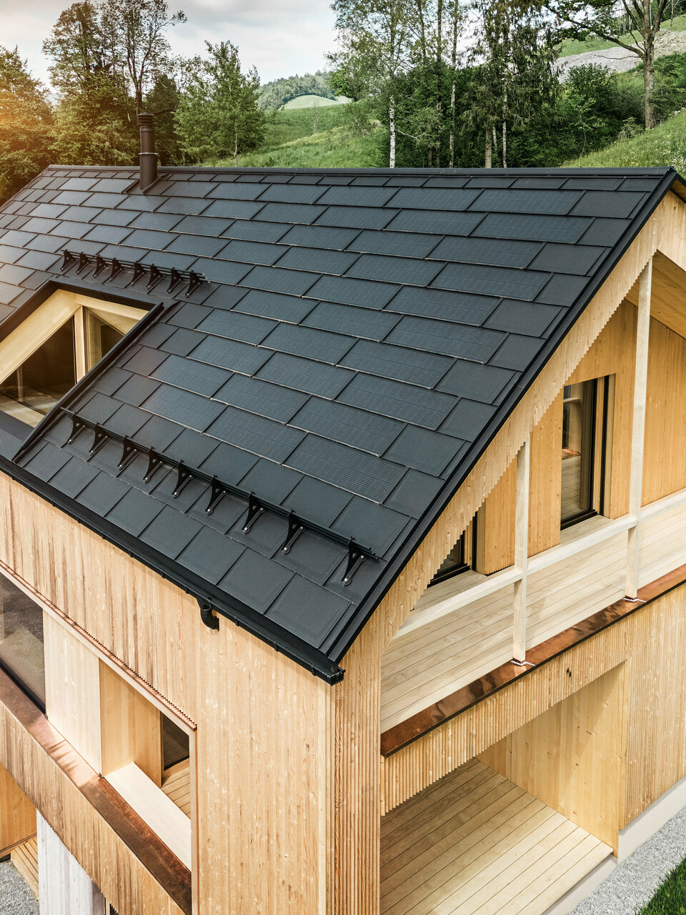 Vista di una moderna casa sostenibile in legno a Egg con un tetto PREFA Solar che si distingue per il suo aspetto scuro, strutturato ed elegante. Le tegole fotovoltaiche sono posizionati strategicamente trale classiche tegole del tetto R.16 per massimizzare il guadagno solare e allo stesso tempo armonizzarsi esteticamente con il design complessivo. La casa è in armonia con il verde circostante, mentre il caldo sole serale enfatizza la texture del legno naturale della facciata e sottolinea la solidità della costruzione. Un esempio armonioso di costruzione attenta all'ambiente e all'efficienza energetica.