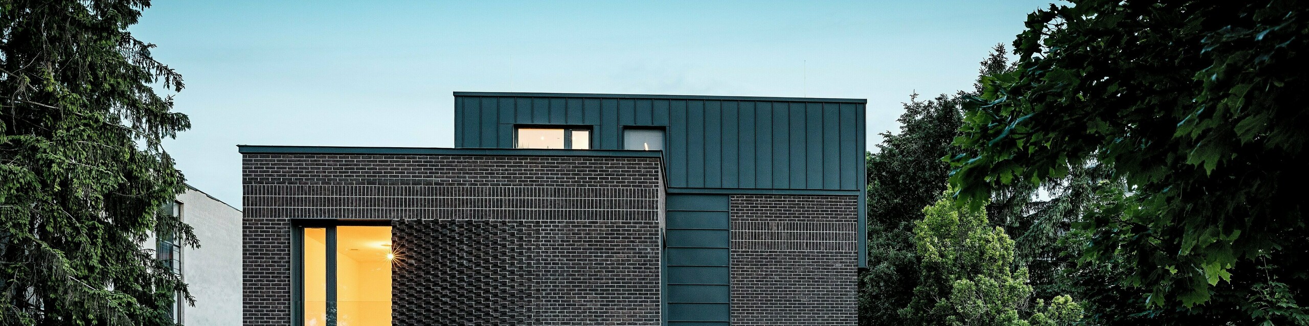 Das Bild zeigt ein modernes Mehrfamilienhaus in Budapest, dessen kubischer Dachaufbau mit PREFALZ Aluminium in P.10 Anthrazit einen markanten Kontrast zur historischen Umgebung bildet. Die vertikalen Winkelstehfalze erzeugen ein spannendes Lichtspiel und betonen die klare Architektursprache. Die hochwertigen PREFA Aluminiumprodukte verleihen dem Gebäude nicht nur eine elegante, zeitlose Optik, sondern überzeugen durch Langlebigkeit, Witterungsbeständigkeit und präzise Verarbeitung. Der anthrazitfarbene Fassaden- und Dachbereich fungiert als kraftvoller Eyecatcher und unterstreicht den innovativen Charakter des Bauwerks.