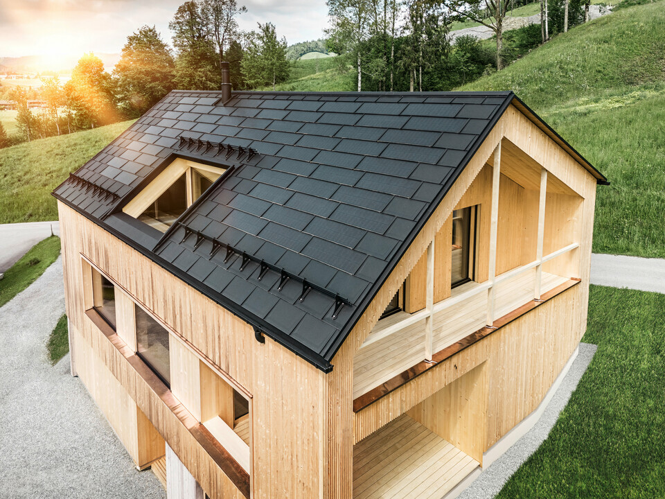 Vista di una moderna casa sostenibile in legno a Egg con un tetto PREFA Solar che si distingue per il suo aspetto scuro, strutturato ed elegante. Le tegole fotovoltaiche sono posizionati strategicamente trale classiche tegole del tetto R.16 per massimizzare il guadagno solare e allo stesso tempo armonizzarsi esteticamente con il design complessivo. La casa è in armonia con il verde circostante, mentre il caldo sole serale enfatizza la texture del legno naturale della facciata e sottolinea la solidità della costruzione. Un esempio armonioso di costruzione attenta all'ambiente e all'efficienza energetica.