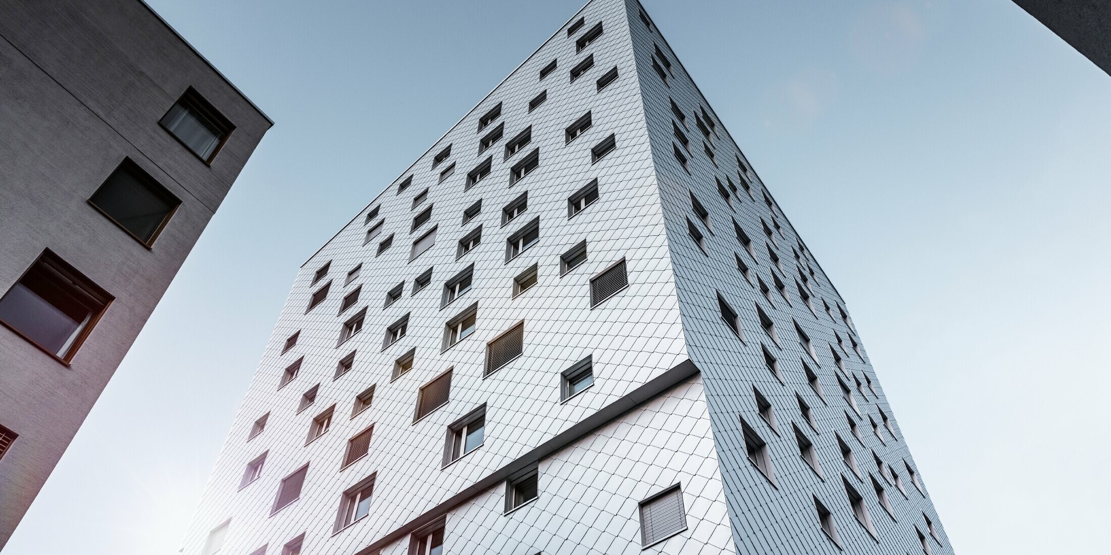 Das Bild zeigt eine Perspektive von unten auf das Solitaire-Hochhaus in Horw, Luzern, das von der Architektin Tilla Theus entworfen wurde. Die Fassade des Gebäudes ist mit eloxierten Aluminiumrauten von PREFA verkleidet, die ein diagonales Muster bilden und die Umgebung reflektieren. Die Fenster sind unregelmäßig angeordnet, was zu der charakteristischen Optik des Gebäudes beiträgt. Im Hintergrund sind Teile anderer Gebäude sichtbar, und der Himmel ist klar und blau.