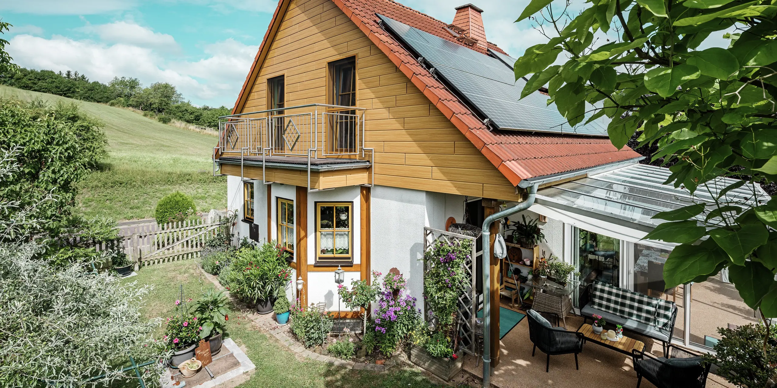 L'immagine ritrae una caratteristica casa unifamiliare a Blieskastel, di cui una parte della facciata è stata rivestita con la Doga PREFA in colore rovere naturale. L'effetto legno dell'alluminio si integra perfettamente con la copertura in laterizio rosso e l'ampio giardino d'inverno. La vegetazione rigogliosa e le aiuole curate incorniciano l'edificio in un contesto armonioso. L'intervento di riqualificazione con i sistemi PREFA conferisce all'edificio un aspetto naturale, garantendo al contempo resistenza agli agenti atmosferici, stabilità cromatica e una durabilità esente da manutenzione.