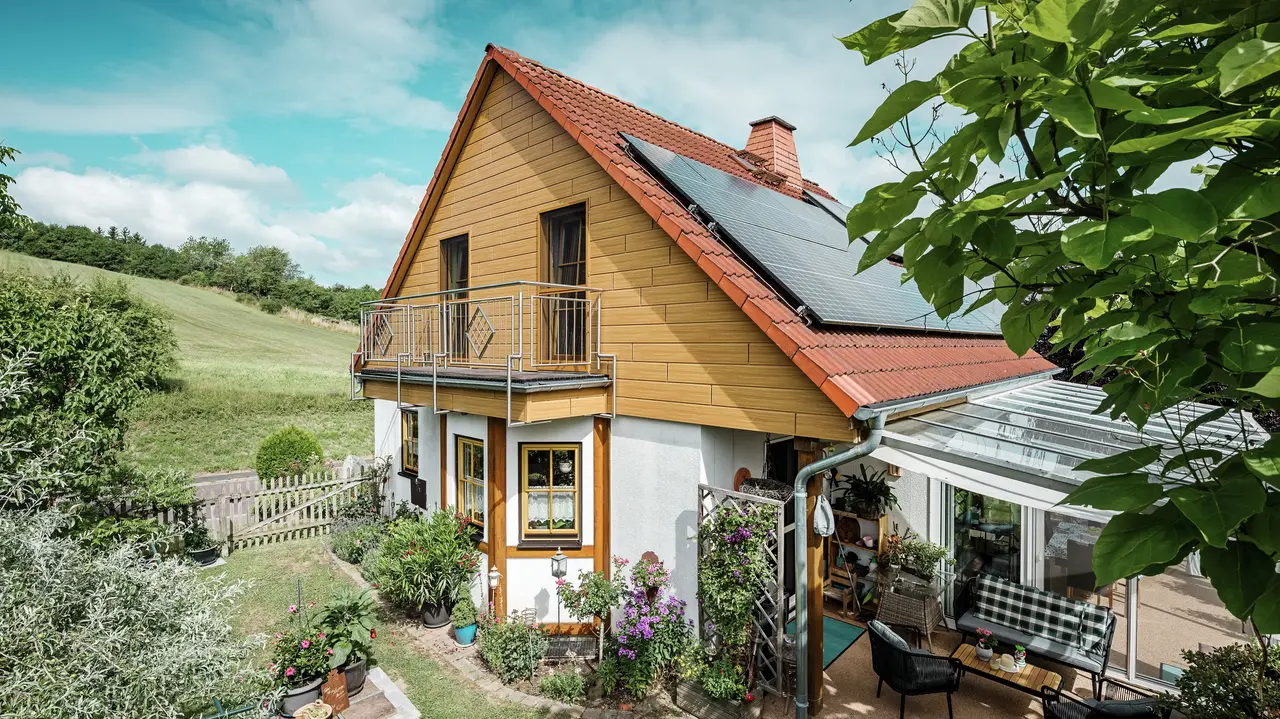 L'immagine ritrae una caratteristica casa unifamiliare a Blieskastel, di cui una parte della facciata è stata rivestita con la Doga PREFA in colore rovere naturale. L'effetto legno dell'alluminio si integra perfettamente con la copertura in laterizio rosso e l'ampio giardino d'inverno. La vegetazione rigogliosa e le aiuole curate incorniciano l'edificio in un contesto armonioso. L'intervento di riqualificazione con i sistemi PREFA conferisce all'edificio un aspetto naturale, garantendo al contempo resistenza agli agenti atmosferici, stabilità cromatica e una durabilità esente da manutenzione.