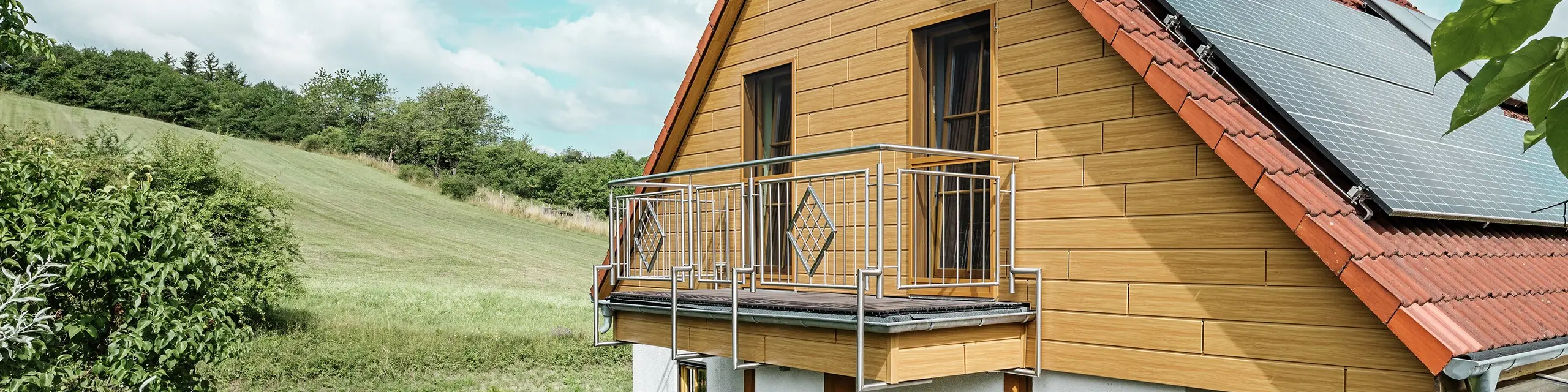 L'immagine ritrae una caratteristica casa unifamiliare a Blieskastel, di cui una parte della facciata è stata rivestita con la Doga PREFA in colore rovere naturale. L'effetto legno dell'alluminio si integra perfettamente con la copertura in laterizio rosso e l'ampio giardino d'inverno. La vegetazione rigogliosa e le aiuole curate incorniciano l'edificio in un contesto armonioso. L'intervento di riqualificazione con i sistemi PREFA conferisce all'edificio un aspetto naturale, garantendo al contempo resistenza agli agenti atmosferici, stabilità cromatica e una durabilità esente da manutenzione.