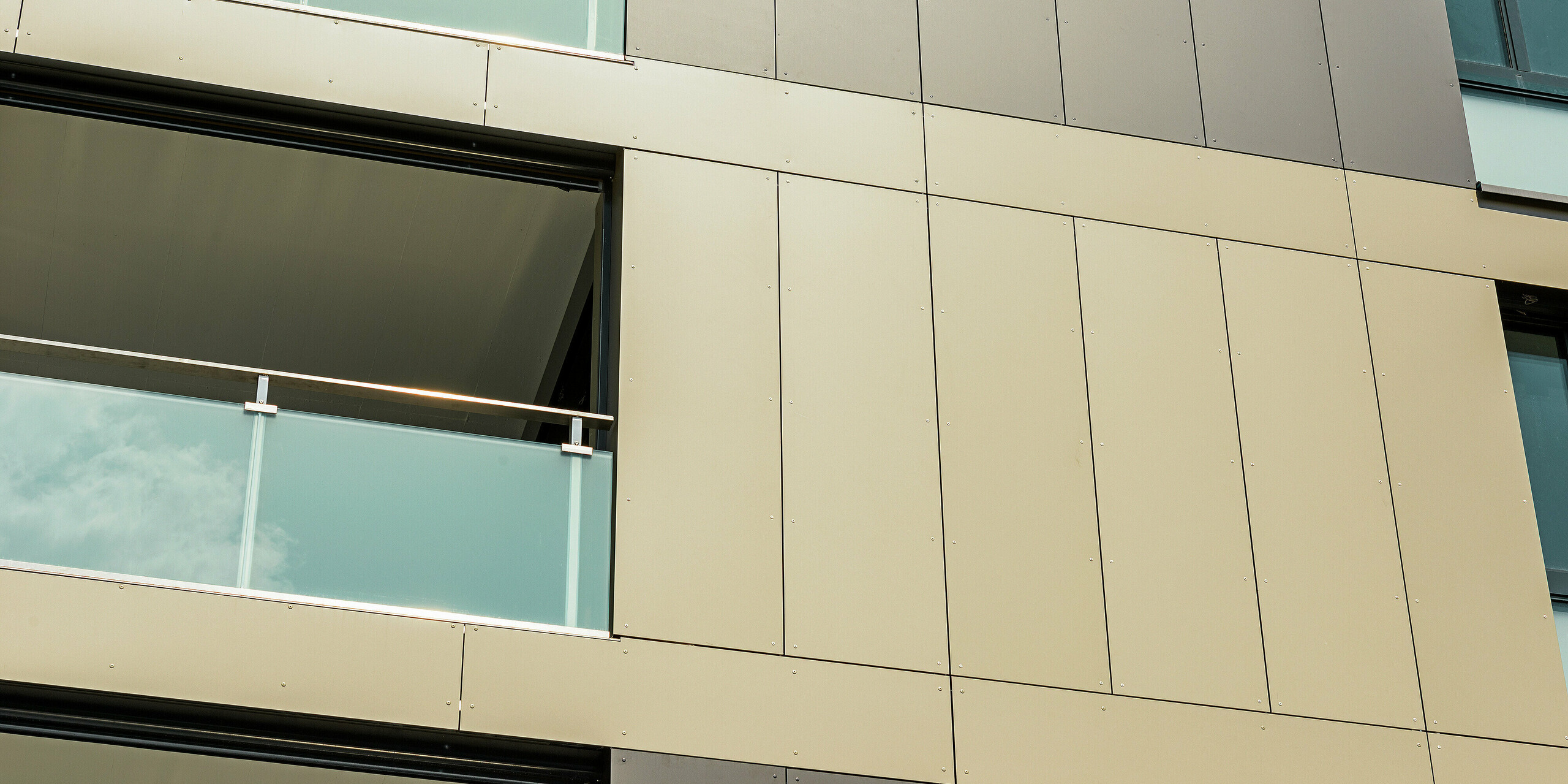 Nahaufnahme der Aluminiumfassade an der kubischen Wohnanlage in Murten (CH). Die Aluminium Verbundplatten von PREFA garantieren eine hohe Widerstandsfähigkeit und Langlebigkeit. Die harmonische Farbgestaltung in Bronze und Schwarzgrau fügt sich perfekt in die Umgebung ein und schützt das Gebäude effektiv vor äußeren Einflüssen wie Wind und Wetter. An den Bildrändern sind Balkone mit Glasverkleidungen zu sehen.
