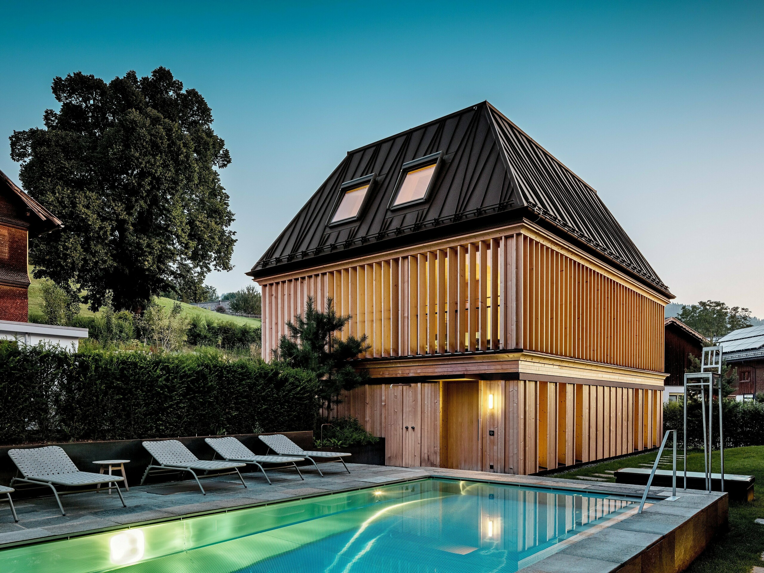 Das „Badehaus“ des Hotel Hirschen in Schwarzenberg in der Dämmerung, mit Blick auf den beleuchteten Außenpool im Vordergrund. Das Gebäude beeindruckt durch sein modernes Walmdach aus PREFALZ in der Farbe P.10 Nussbraun mit präzise ausgeführten, teils schräg verlaufenden Stehfalzen. Zwei Dachfenster sorgen für Tageslicht im Inneren. Die Fassade besteht aus vertikalen, geschoßhohen Holzlamellen, die dem Baukörper Struktur und Leichtigkeit verleihen. Im unteren Bereich dominiert naturbelassenes Holz. Umgeben von gepflegtem Grün und eingebettet in die Hügellandschaft des Bregenzerwaldes entsteht eine harmonische Verbindung aus Architektur, Natur und Erholung.