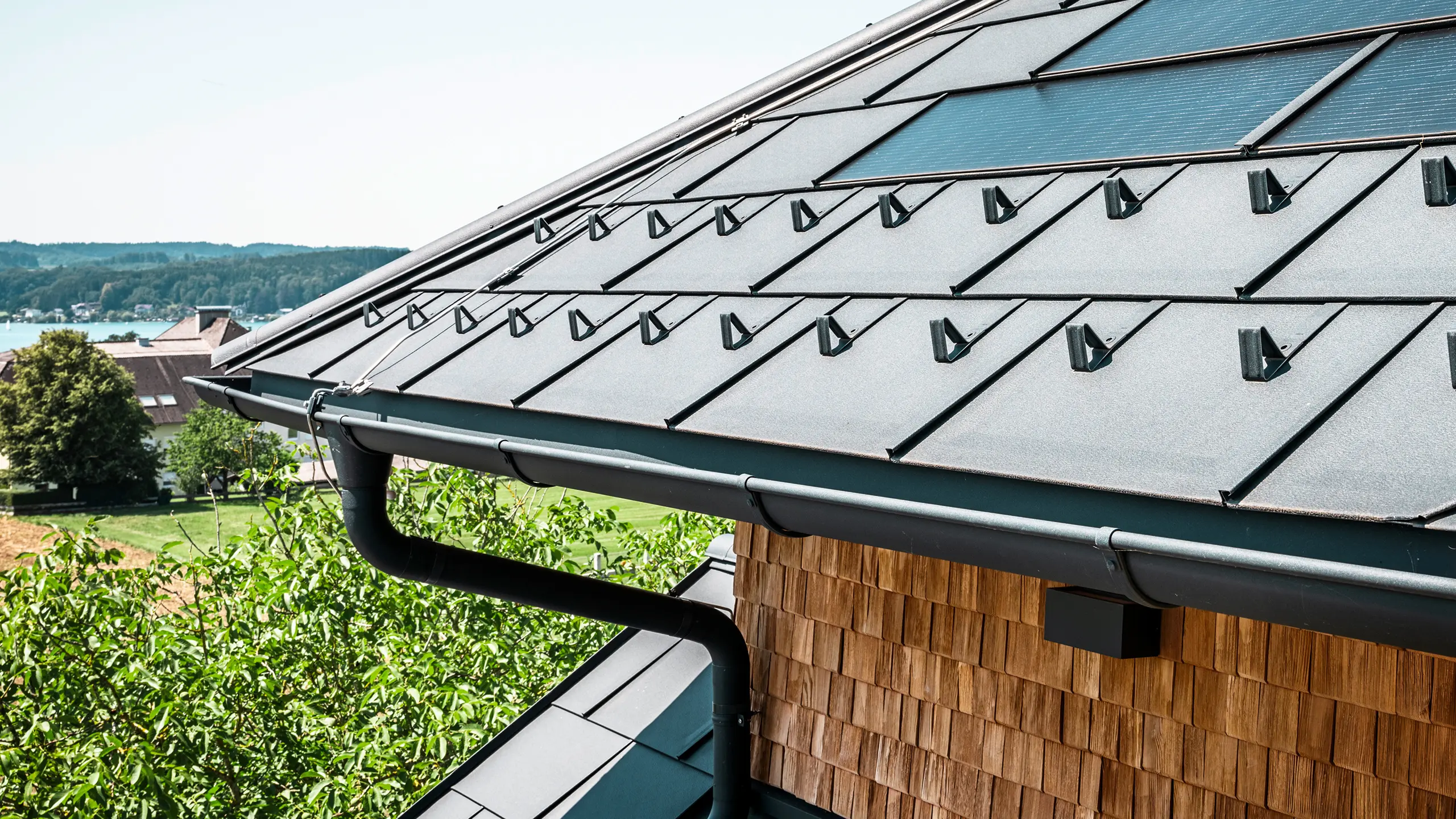 Die Zenz'n Stub'n in Schörfling am Attersee präsentiert ein neu saniertes Dach mit innovativen PREFA Solardachplatten und langlebigen PREFA Dachplatten R.16 in P.10 Anthrazit. Das stilvolle Aluminiumdach verbindet modernes Design mit nachhaltiger Energiegewinnung. Auch die Dachentwässerung, bestehend aus Ablaufrohr und Hängerinne in P.10 Anthrazit, fügt sich harmonisch in das Gesamtbild ein und unterstreicht die hochwertige Verarbeitung. Im Hintergrund blitzt das Blau des Attersees hervor.