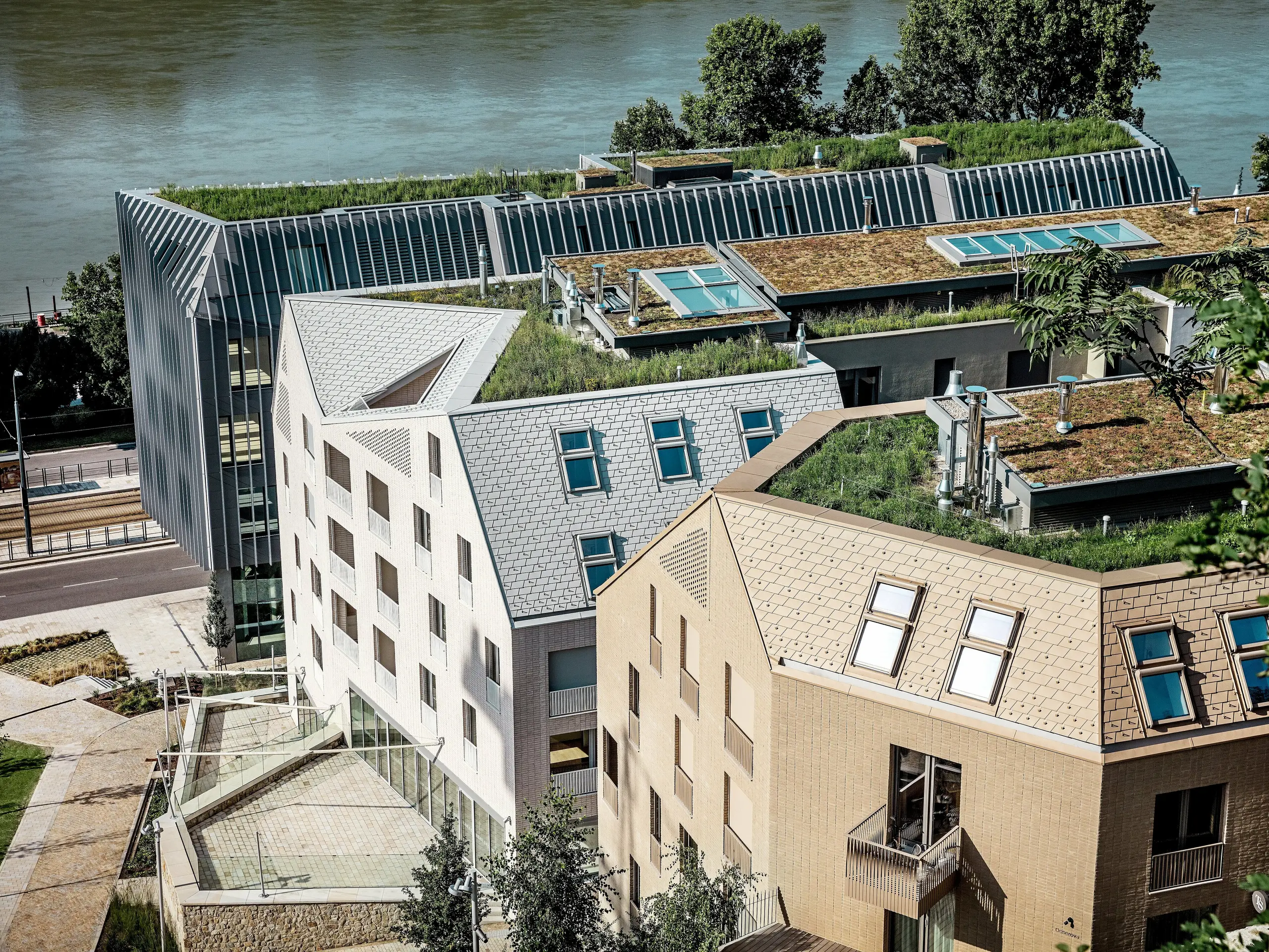 Modernes Stadtquartier in Bratislava mit markanter Architektur und begrünten Dachlandschaften direkt am Flussufer: Die filigranen, steil geneigten Dächer und Gauben werden von hochwertigen PREFA Aluminium Dachschindeln in Prefaweiß, Sandbraun und Bronze geprägt und ziehen sofort alle Blicke auf sich. Großzügige Dachflächenfenster, klare Linien und üppige Dachbegrünung verbinden Design und Nachhaltigkeit. Die langlebigen, wartungsarmen PREFA Aluminiumlösungen überzeugen durch Widerstandskraft, Leichtigkeit und Witterungsbeständigkeit – ideal für anspruchsvolle urbane Bauprojekte.