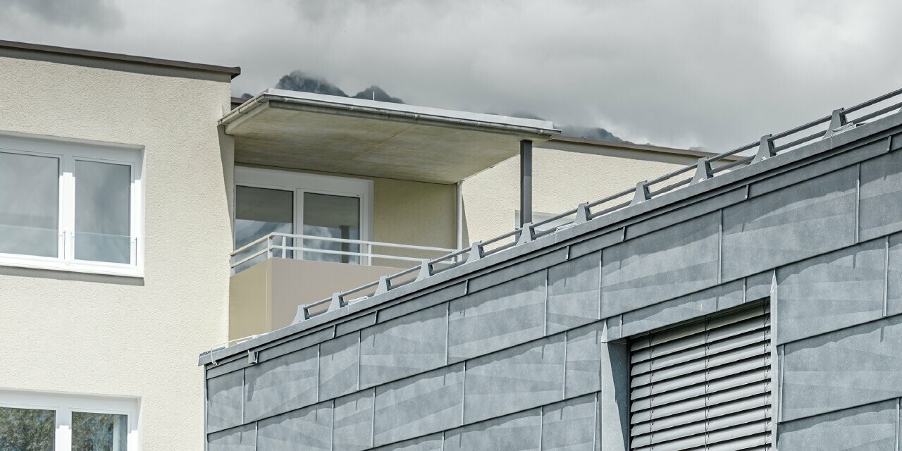 Surface de façade avec deux fenêtres ; la façade est habillée avec le panneau de façade FX.12 PREFA couleur gris pierre.