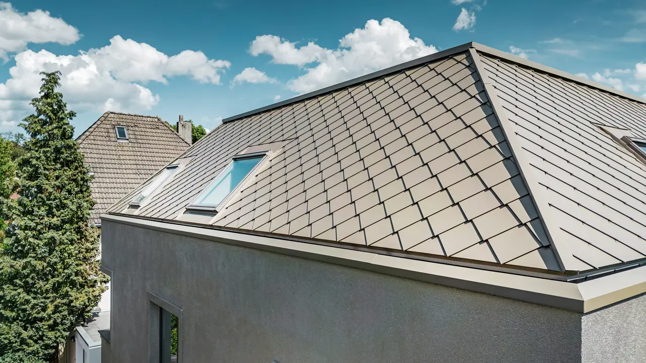 Primo piano del tetto a falde di una moderna casa unifamiliare con rivestimento in alluminio color bronzo di PREFA. La costruzione del tetto presenta una finitura di alta qualità con lucernari integrati che forniscono ulteriore luce naturale all'interno. Le linee chiare e la superficie lucida delle tegole in alluminio sottolineano il design moderno ed elegante della casa. 