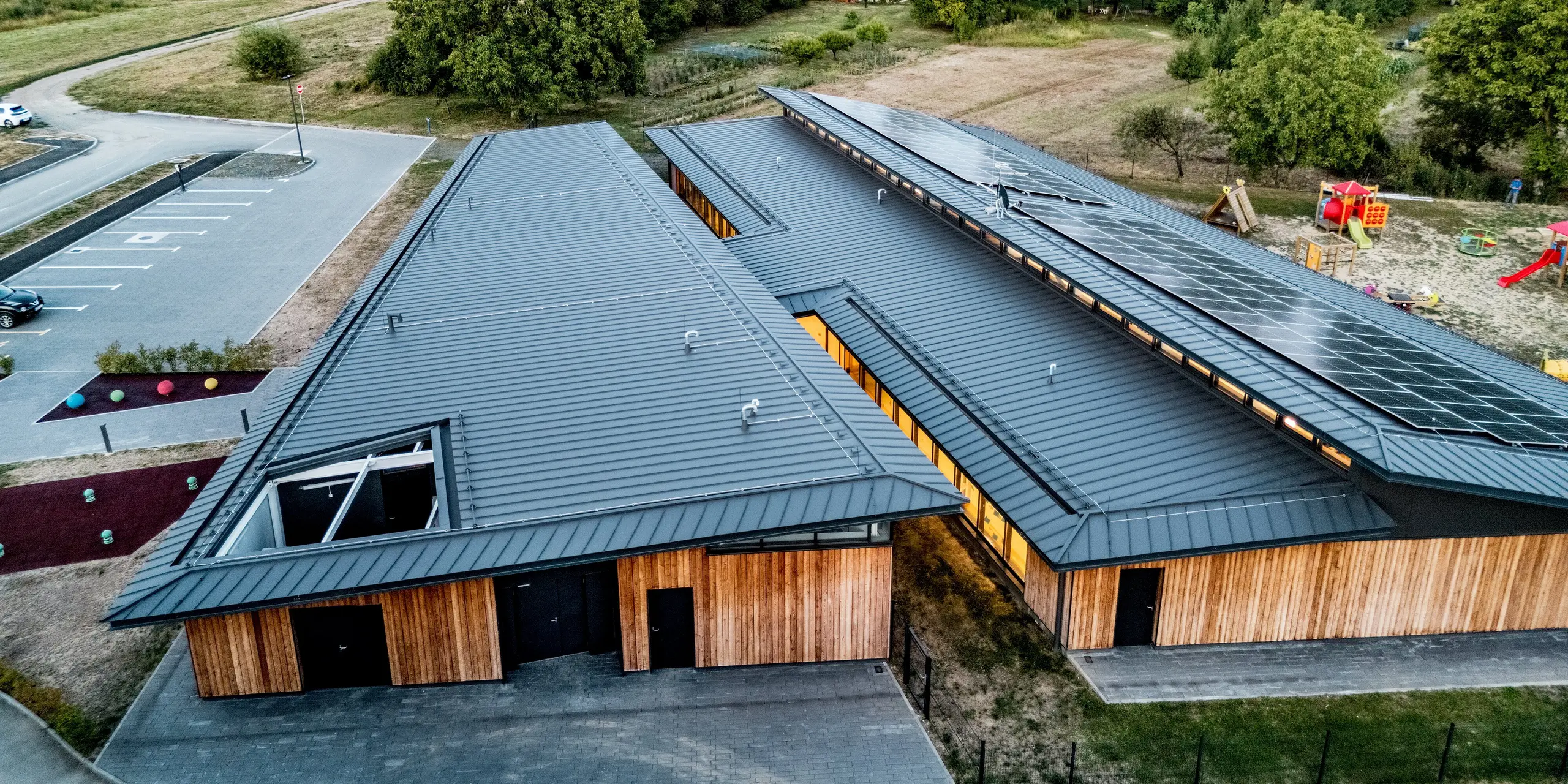 Luftaufnahme des Kindergartens Svarozic in Kroatien mit klar gegliederter Dachlandschaft, Holzfassade und weitläufigem Außengelände. Die anthrazitfarbene PREFALZ Dachdeckung aus Aluminium prägt das moderne Erscheinungsbild und verbindet langlebigen Witterungsschutz mit eleganter Optik. Umgeben von Grünflächen und Spielbereichen zeigt sich die harmonische Einbettung des Gebäudes in die Umgebung.