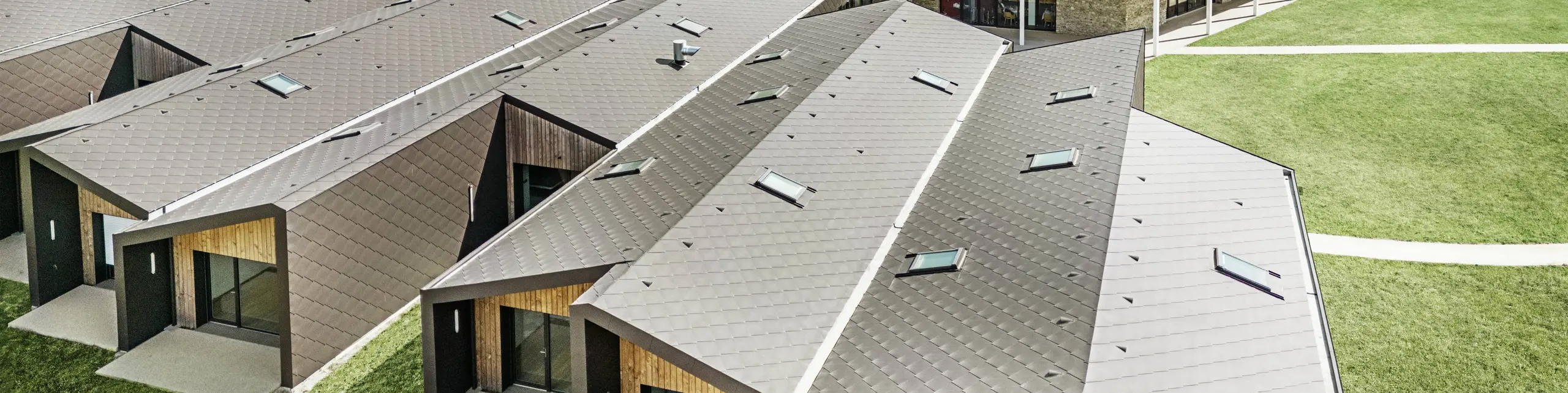 L'immagine mostra una veduta aerea di un moderno complesso edilizio rivestito con scaglie 44 x 44 di PREFA per tetti e facciata, nel colore marrone testa di moro. Il paesaggio del tetto è caratterizzato da forme geometriche combinate con grandi facciate in vetro ed elementi in legno. Sullo sfondo si intravede un'antica chiesa che forma un contrasto armonioso con l'architettura moderna. Il prato verde e la natura circostante sottolineano il design aperto e sostenibile dell'intero complesso.