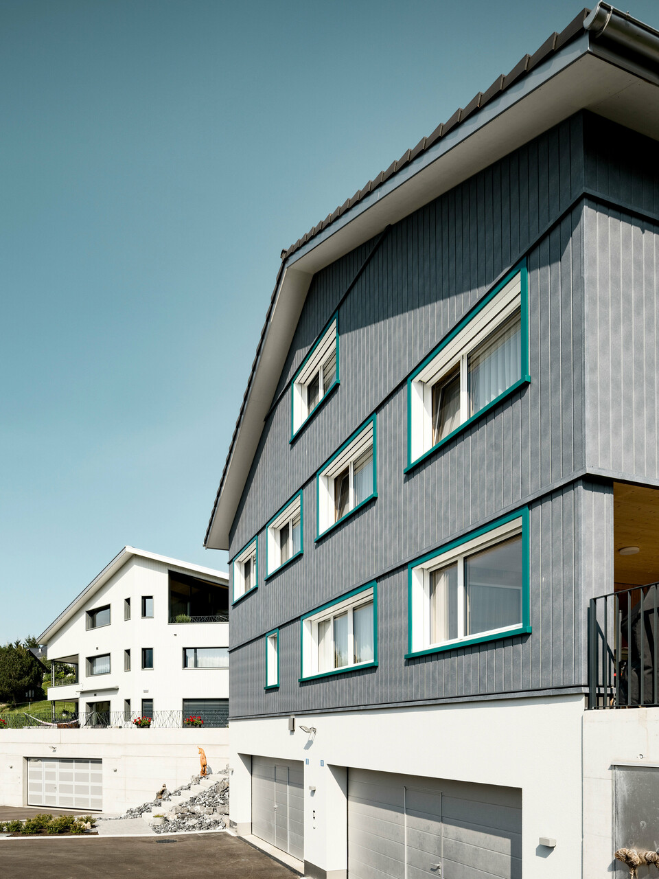 Ansicht zweier benachbarter Mehrfamilienhäuser in Weissbad, Schweiz, wobei das vordere Gebäude mit seiner PREFA Siding Fassade in P.10 Steingrau einen starken visuellen Eindruck vermittelt. Die vertikal montierten Sidings schaffen eine elegante und schützende Hülle, während die türkisen Fensterrahmen farbliche Akzente setzen. Das Bild fängt das Zusammenspiel der modernen Materialien mit der traditionellen Architektur ein und demonstriert die vielseitige Einsetzbarkeit von PREFA Aluminiumprodukten.