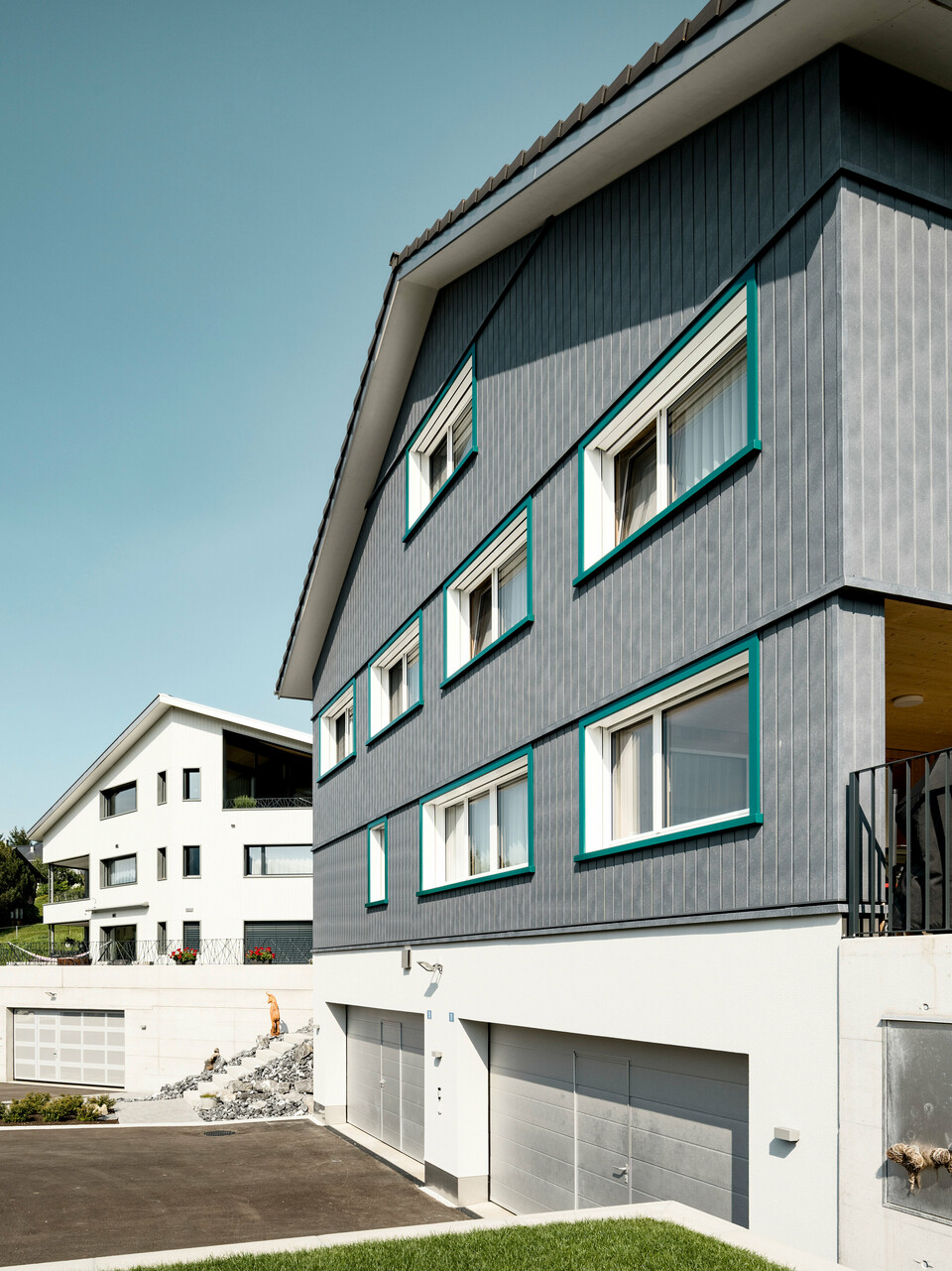 Ansicht zweier benachbarter Mehrfamilienhäuser in Weissbad, Schweiz, wobei das vordere Gebäude mit seiner PREFA Siding Fassade in P.10 Steingrau einen starken visuellen Eindruck vermittelt. Die vertikal montierten Sidings schaffen eine elegante und schützende Hülle, während die türkisen Fensterrahmen farbliche Akzente setzen. Das Bild fängt das Zusammenspiel der modernen Materialien mit der traditionellen Architektur ein und demonstriert die vielseitige Einsetzbarkeit von PREFA Aluminiumprodukten.