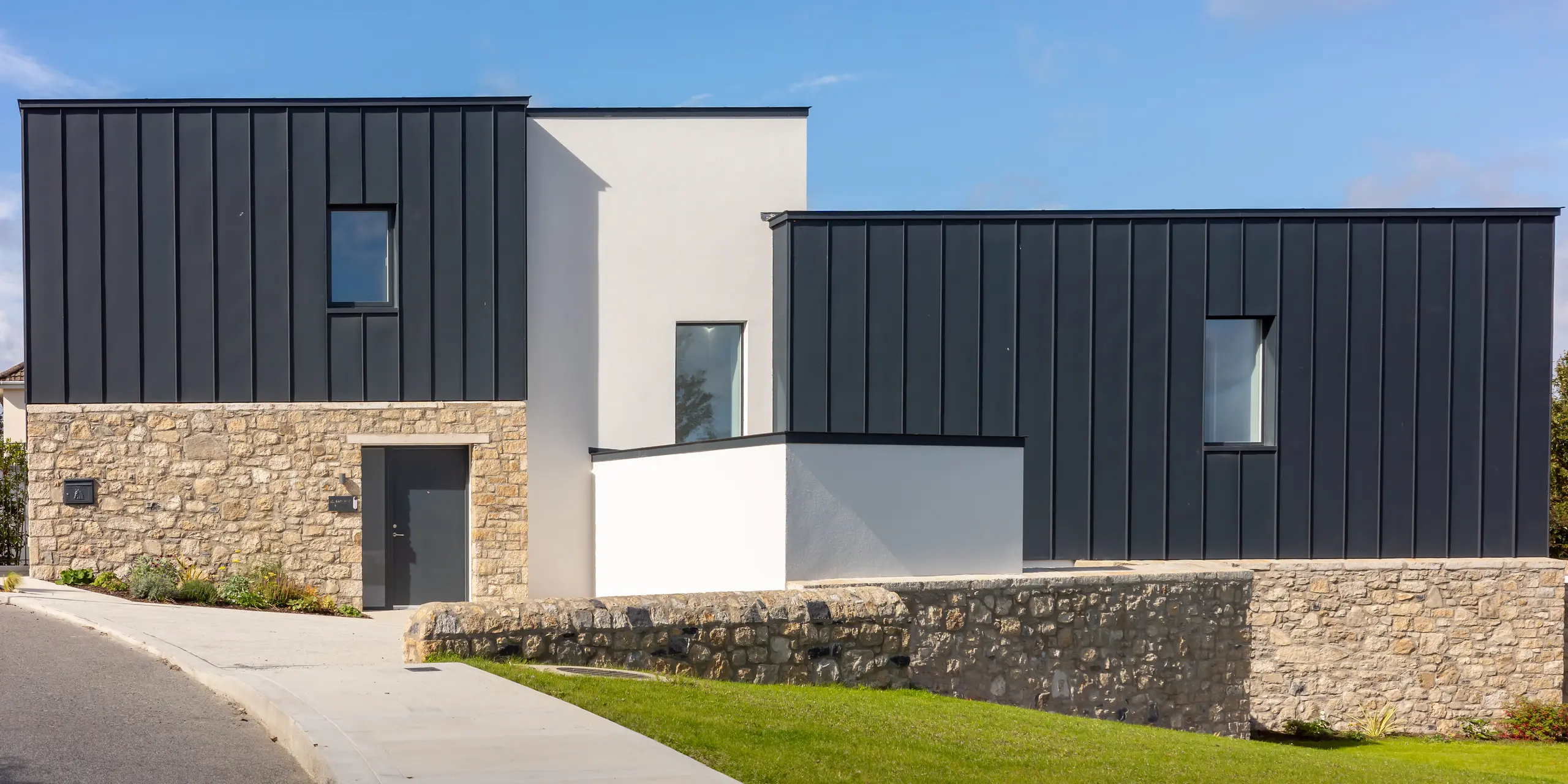 Immeuble résidentiel moderne à Watson Place, aux lignes épurées, murs en pierre naturelle et remarquable toiture et façade en PREFALZ P.10 anthracite. Les surfaces lisses en aluminium créent d'élégants contrastes avec la pierre et le plâtre, tout en assurant une protection durable contre les intempéries. Le jeu des matériaux confère au bâtiment une allure intemporelle et raffinée.