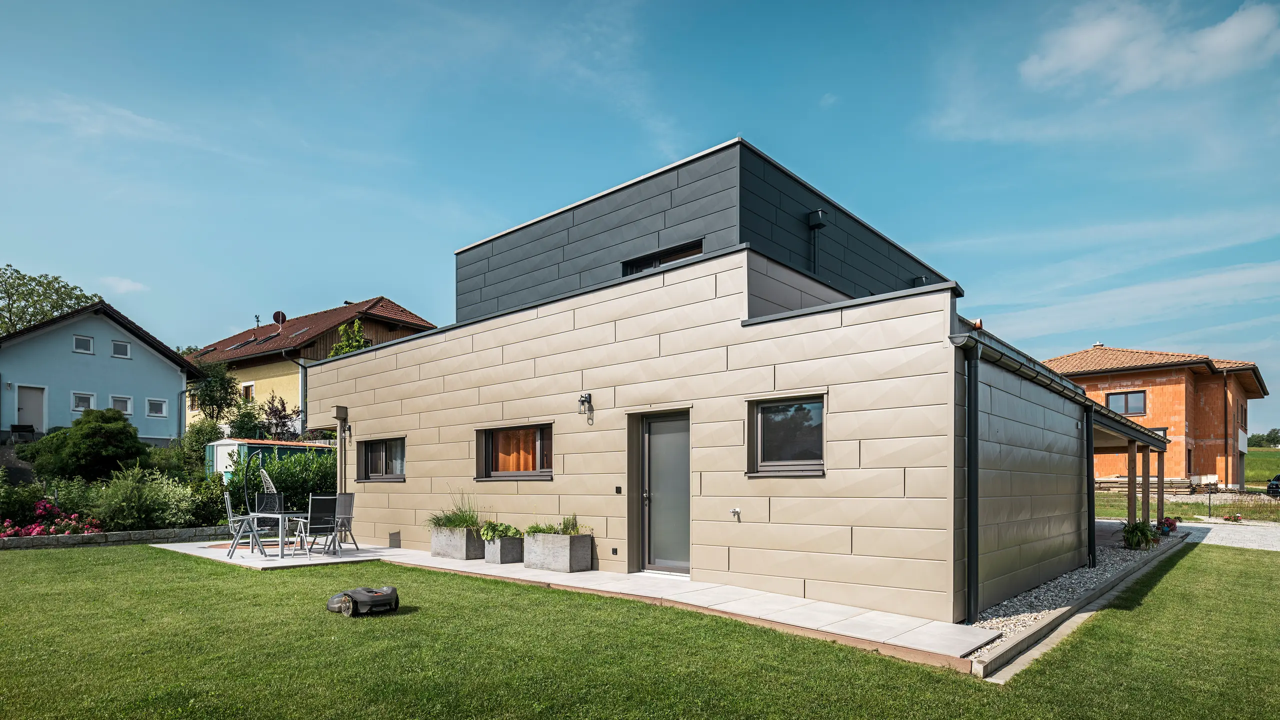 Maison individuelle moderne à Wippenham, en Autriche, avec façade en aluminium PREFA bicolore bronze et anthracite. La façade est conçue avec des panneaux de façade Siding.X, qui assurent un look élégant et contemporain. Le système de gouttière PREFA intégré avec collecteur d'eau et tuyau d'évacuation ainsi qu'un jardin bien entretenu avec terrasse soulignent le design moderne de la maison.