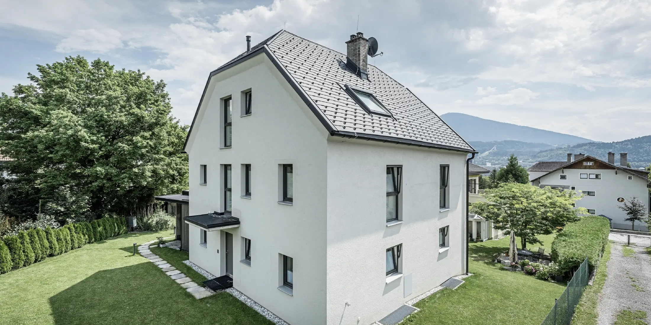 Maison individuelle moderne de trois étages avec une façade claire en crépi et un toit en pente gris sombre en aluminium PREFA. Le bâtiment dispose de plusieurs fenêtres étroites et de gouttières anthracite. Il est situé dans un jardin bien entretenu avec une pelouse, des arbres et des haies. À l'arrière-plan, on aperçoit d'autres maisons et un paysage montagneux.