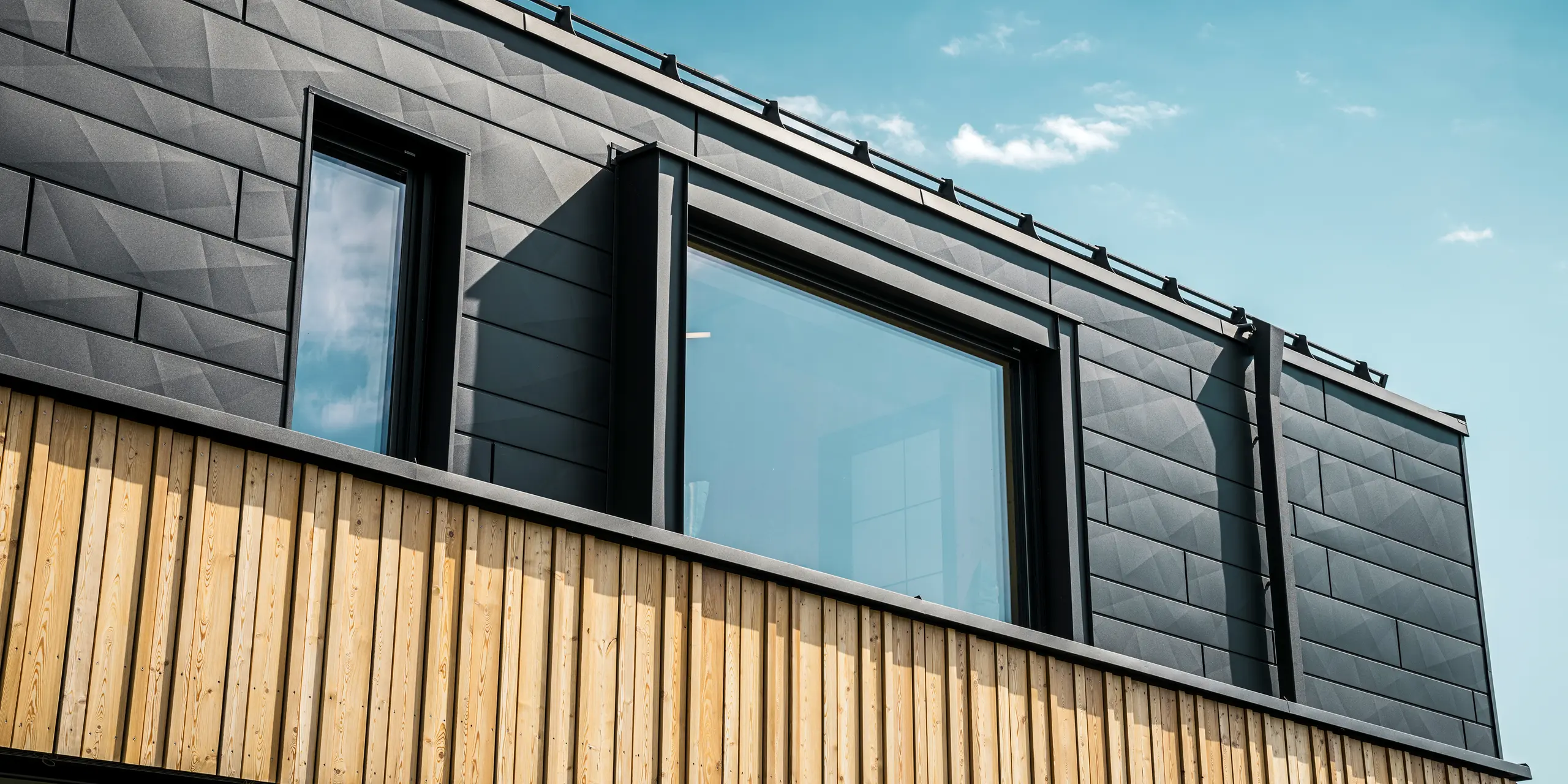 Gros plan sur l'étage supérieur de la maison témoin Autark dans le centre de maisons préfabriquées Blaue Lagune, à Vösendorf (AT). La façade est composée de panneaux en aluminium PREFA Siding.X en P.10 noir qui se distinguent par leur aspect particulier avec différents plis. En dessous se trouve un habillage en bois élégant qui forme un contraste harmonieux avec l'aluminium foncé. De grandes fenêtres laissent entrer beaucoup de lumière à l'intérieur du bâtiment et soulignent le design moderne.