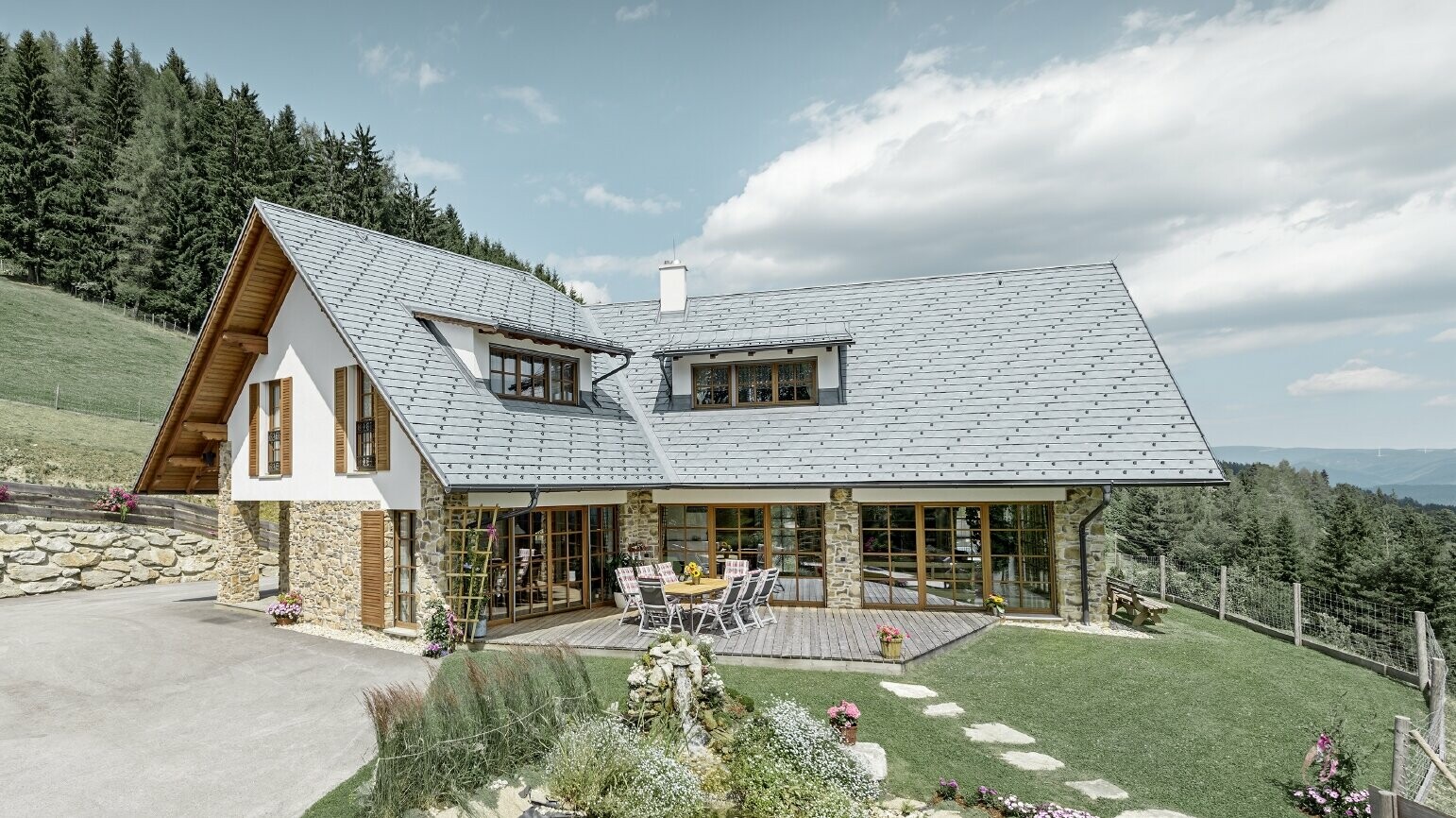 Construite en forme de L et à flanc de coteau, cette maison individuelle présente un toit à deux pans avec deux lucarnes rampantes. La toiture a été recouverte de bardeaux de toiture PREFA couleur gris pierre. Un étang se trouve au premier plan et on peut voir une forêt en arrière plan.