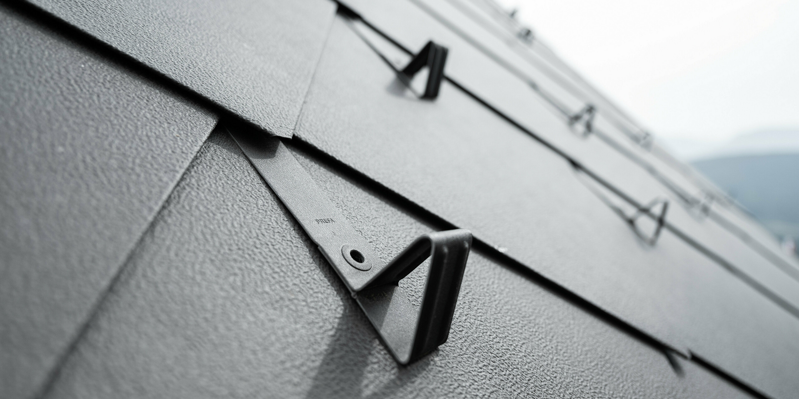 Detailaufnahme eines Aluminiumdaches aus PREFA Dachraut 44x44 in der Farbe P.10 Braun, ausgestattet mit einem funktionalen Schneeschutzsystem. Das Bild zeigt die strukturierte Oberfläche der Rauten sowie die präzise Befestigung der Schneeschutzhalterungen, die nahtlos in das Dachdesign integriert sind und für zusätzliche Sicherheit während der Wintermonate sorgen. Die robusten Schneestopper sind gleichmäßig angeordnet und sorgen dadurch für ein harmonisches Gesamtbild.