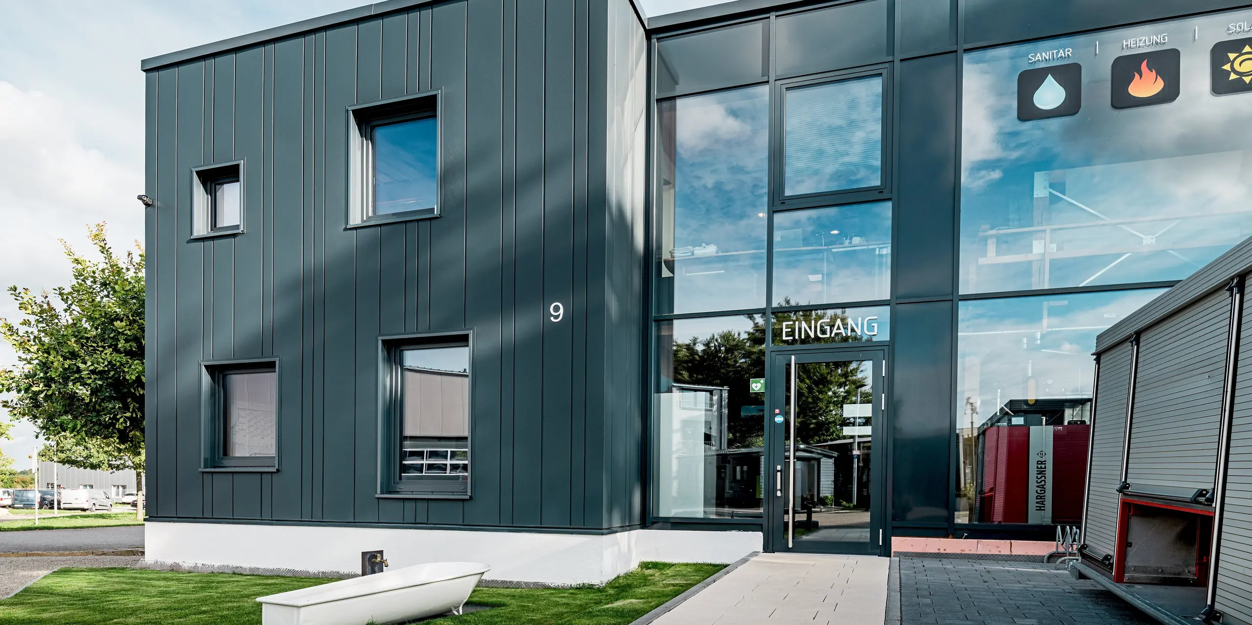 Cette image présente l'entrée moderne de l'entreprise de construction Schädel GmbH, caractérisée par une façade en aluminium PREFA P.10 anthracite du plus bel effet. Les Sidingsverticaux de largeurs variables créent une structure dynamique. Les surfaces vitrées réfléchissantes produisent un jeu de lumière et de matière captivant. Robuste, la façade PREFA allie protection durable et élégance contemporaine.