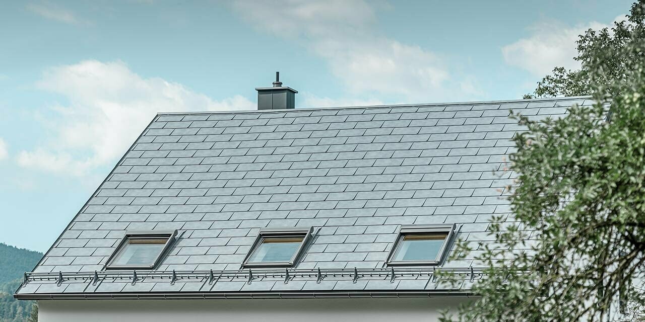 Classico tetto a due falde, rivestito con tegola R.16 PREFA in grigio pietra con tre lucernari e sistema paraneve; la facciata è intonacata di bianco.