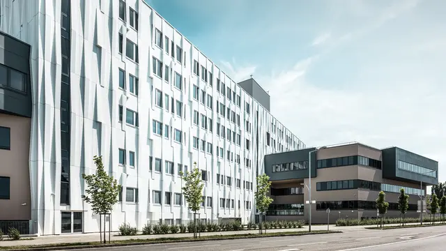 Seitliche Ansicht des Universitätsklinikums in St. Pölten, Niederösterreich. Die robuste Aluminiumfassade macht das Gebäude zu einem Blickfang in der Stadt. Im Vordergrund ist eine Straße mit einigen Bäumen zu sehen.