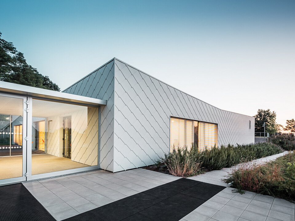 L'asilo nido con la facciata composta da scaglie in bianco PREFA nella città francese di Otterswiller, progettata da Urbane Kultur, dalla prospettiva normale laterale; davanti un marciapiede e lo spazio verde.