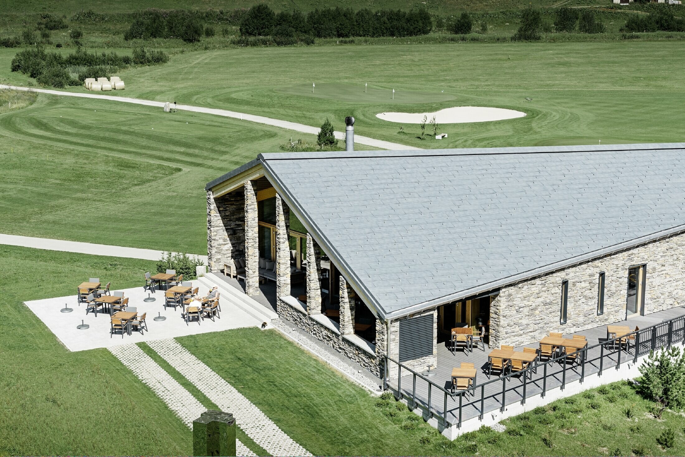 Il moderno punto di ritrovo del campo da golf a Andermatt con una facciata in pietra e il pannello per coperture FX.12 PREFA in alluminio in grigio pietra