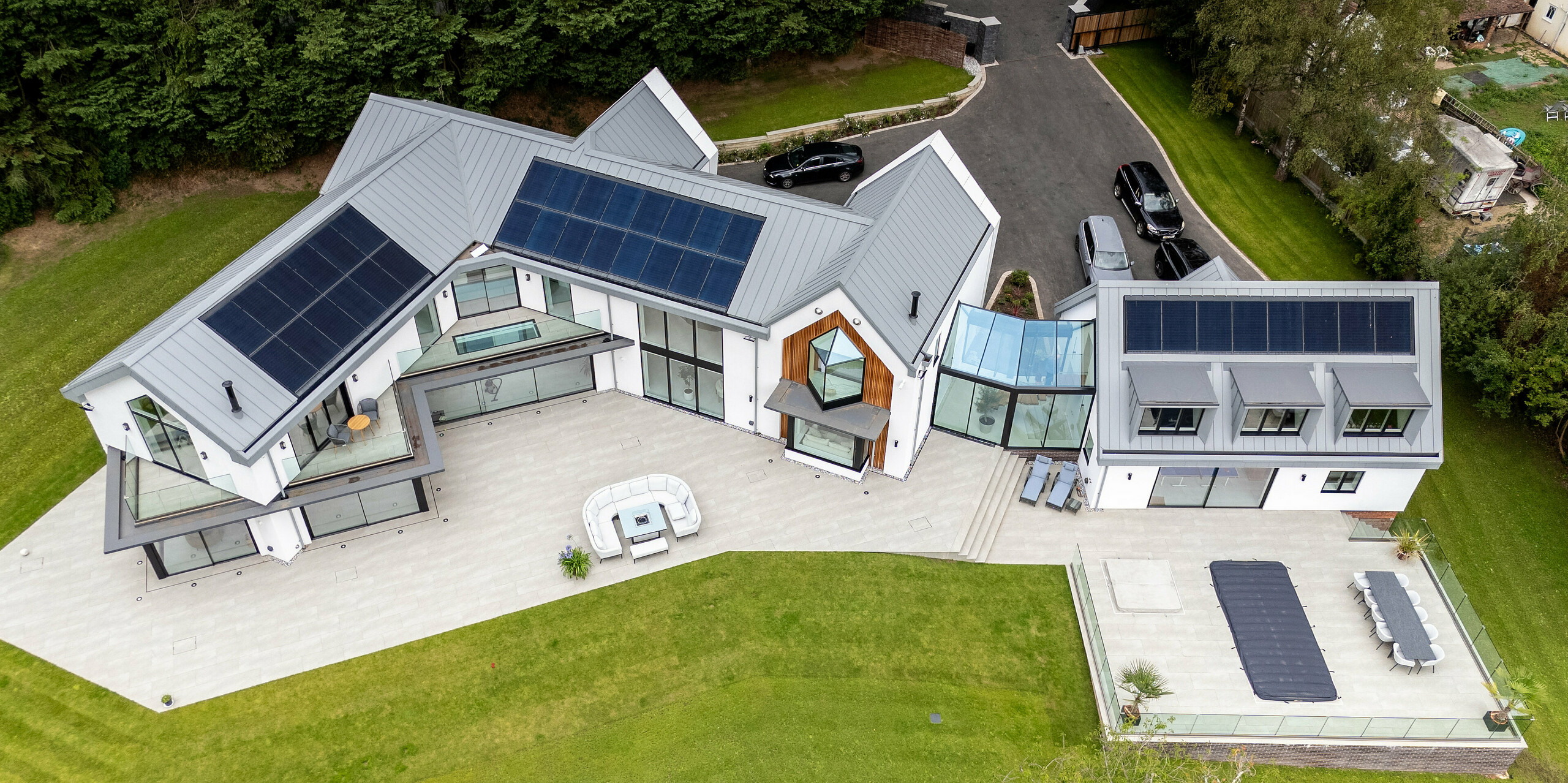 L'image offre une vue aérienne imprenable sur cette villa moderne située à Draycott-in-the-Clay, reconnaissable à sa remarquable toiture PREFALZ gris quartz. La toiture à joints debout s'intègre harmonieusement au paysage verdoyant environnant et souligne le design moderne et élégant de la maison. Les panneaux solaires, parfaitement intégrés à la toiture, témoignent de son orientation écoénergétique. La vaste terrasse et le jardin soigné complètent le tableau de cette architecture innovante. L'utilisation de PREFALZ garantit une qualité et une durabilité optimales.
