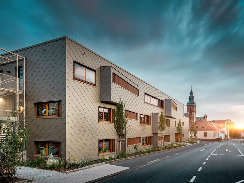 Im sanften Licht des Sonnenuntergangs zeigt sich die Kindertagesstätte in Alzenau von ihrer stimmungsvollsten Seite. Die Fassade aus bronzefarbenen PREFA Wandrauten 44 × 44 reflektiert das Abendlicht und betont die hochwertige, strukturierte Oberfläche des modernen Hybridgebäudes. Die klar gegliederte Architektur mit versetzten Fensterbändern und vertikalen Holzlamellen steht in spannendem Kontrast zur historischen Kirche im Hintergrund. Links im Bild sorgt ein filigranes Metallgerüst für leichte Transparenz beim Außentreppenhaus. Der gepflegte Grünstreifen entlang des Gebäudes unterstreicht den ökologischen Anspruch. Dieses Bild verbindet eindrucksvoll moderne Baukultur, nachhaltige PREFA Aluminiumtechnologie und harmonische Einbettung in das historische Stadtbild von Alzenau.