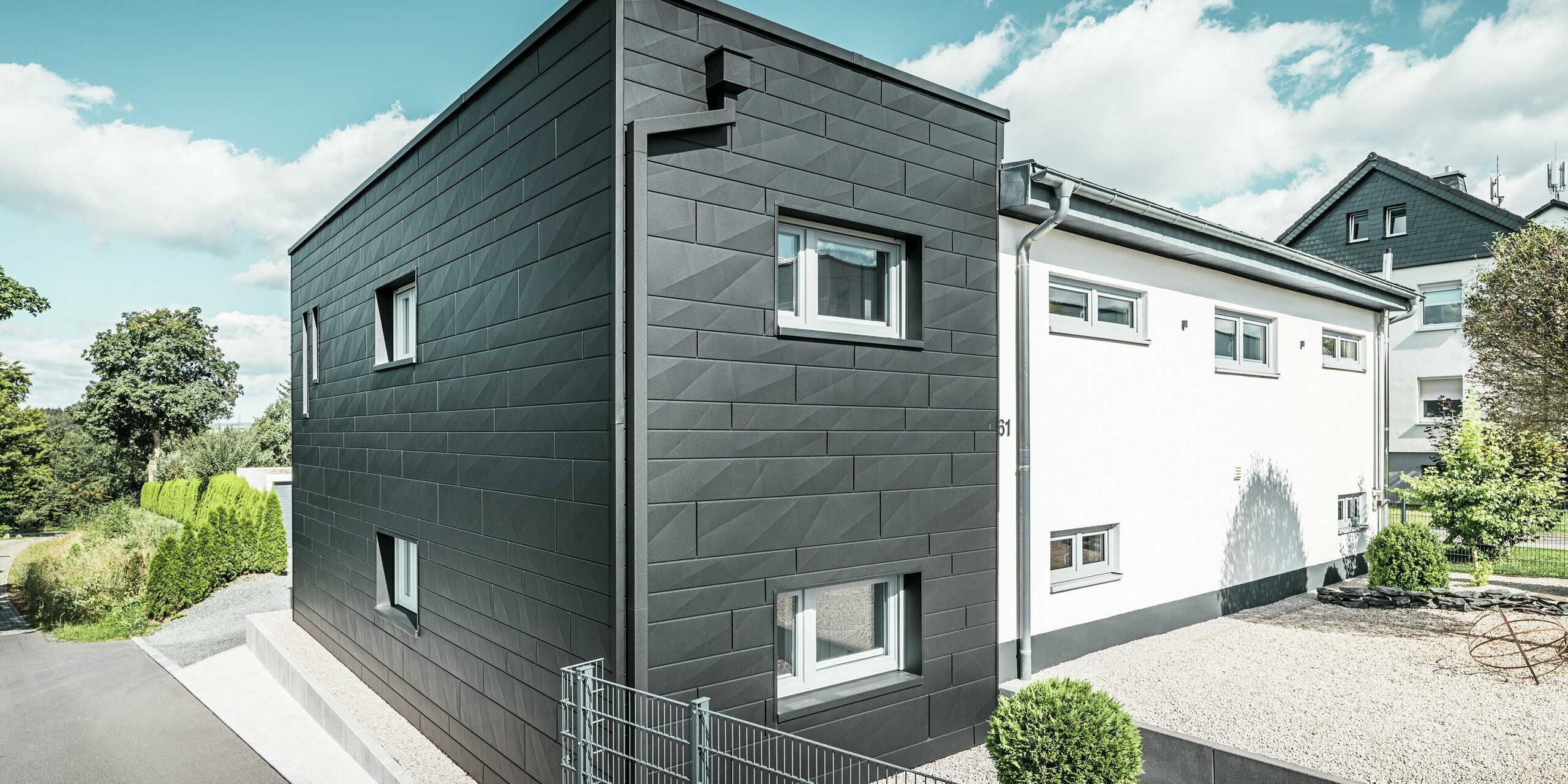 Die markante PREFA Siding.X Fassade in P.10 Schwarz prägt die moderne Architektur des sanierten Einfamilienhauses in Teuschnitz. Die klar strukturierten Aluminiumpaneele verleihen dem Gebäude eine elegante, geometrische Ästhetik. Die integrierte PREFA Dachentwässerung mit dem Quadratrohr fügt sich nahtlos in das Gebäude ein und sorgt für eine funktionale sowie optisch ansprechende Lösung. Der grüne Garten und die Kiesfläche vor dem Haus ergänzen das minimalistische Design und betonen den modernen Charakter des Hauses.