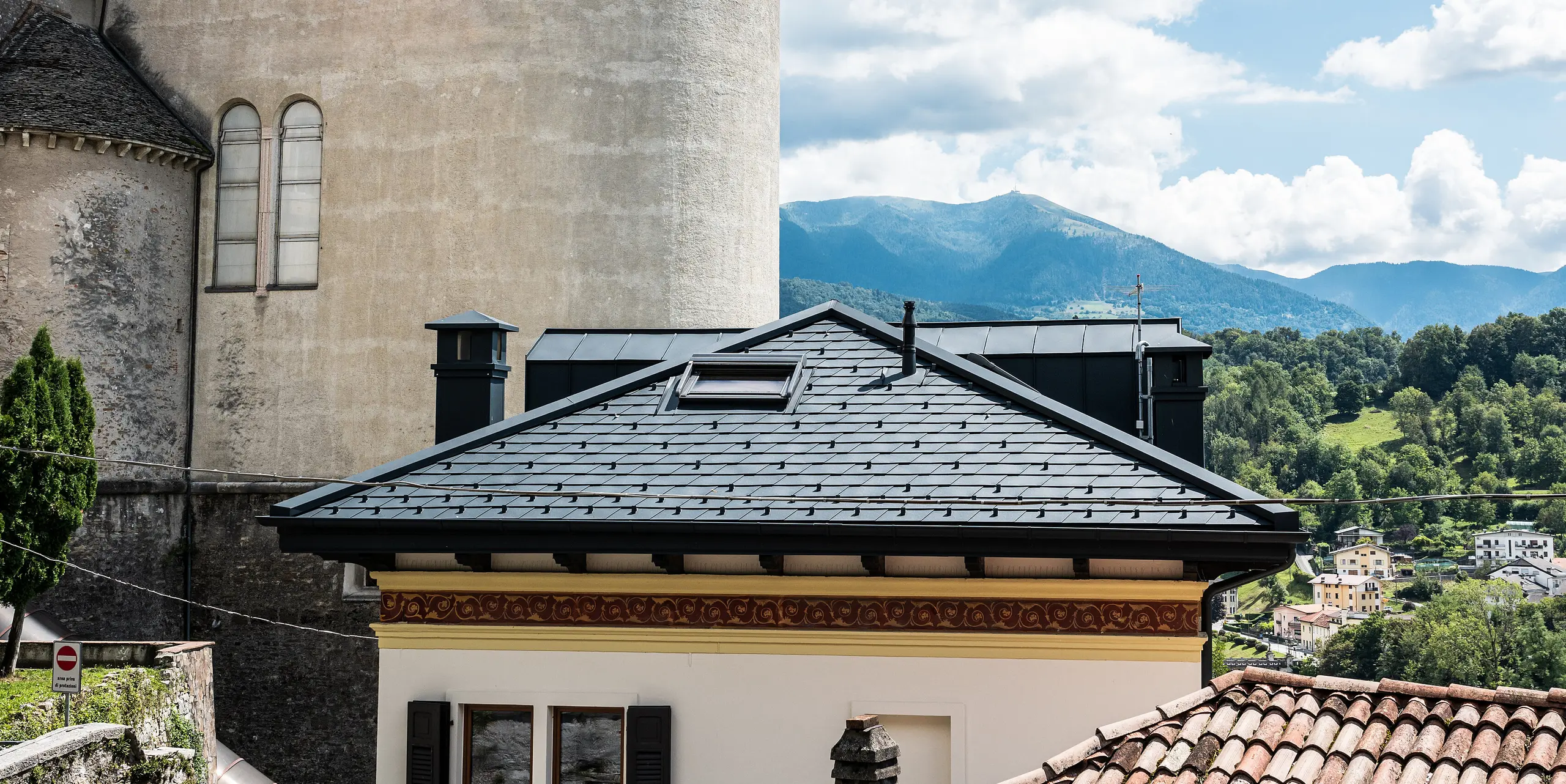 Historische und moderne Architektur vereint in Belluno, Italien, mit einem sorgfältig modernisierten Wohngebäude, dessen Dach mit anthrazitfarbenen PREFA Aluminiumdachschindeln versehen ist. Das Gebäude steht im Einklang mit der umgebenden traditionellen Struktur und dem grünen, hügeligen Hintergrund, während die klare Linienführung und die Kaminverkleidungen das zeitgenössische Design und die innovative Nutzung von langlebigen Materialien hervorheben.