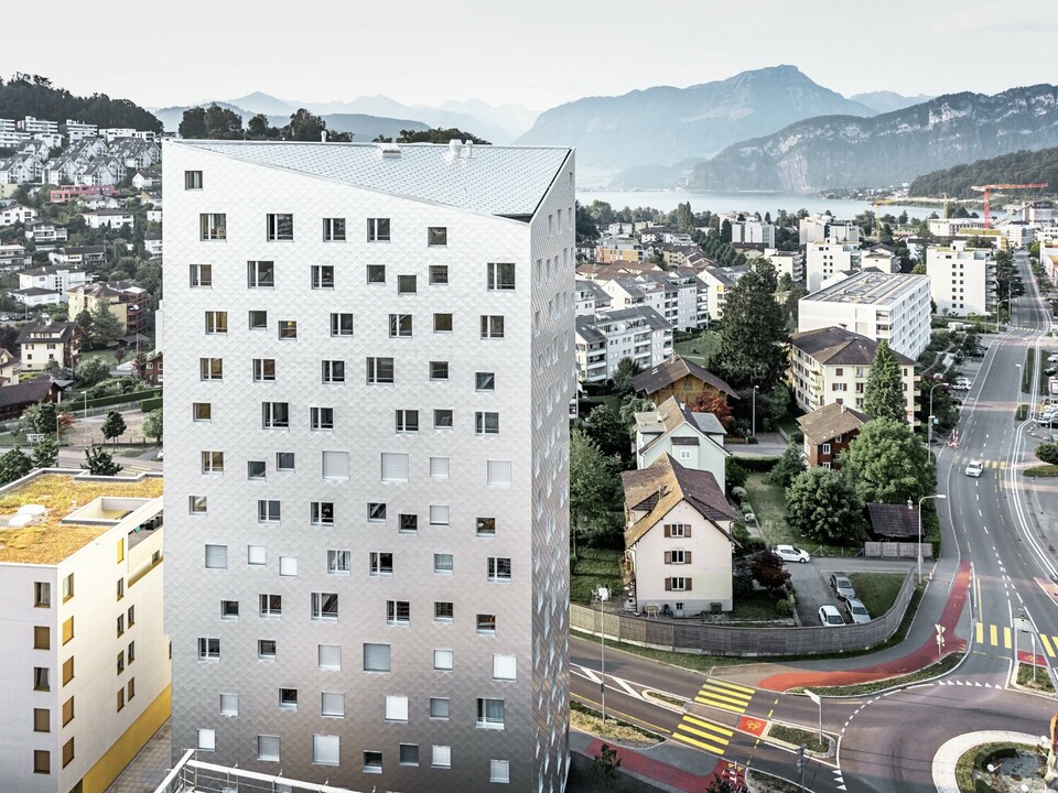 Das Bild zeigt das Solitaire-Hochhaus in Horw, einem Vorort von Luzern, das von der Schweizer Architektin Tilla Theus entworfen wurde. Das Hochhaus hat eine Fassade aus eloxierten Aluminiumrauten, die die Umgebung und die Wolken reflektieren. Die Fenster sind unregelmäßig angeordnet und als 'tanzende Fenster' bekannt. Im Hintergrund sind weitere Gebäude, Straßen und die malerische Berglandschaft der Schweiz zu sehen. Das Hochhaus steht inmitten einer städtischen Umgebung mit einer Mischung aus Wohnhäusern und städtischer Infrastruktur.