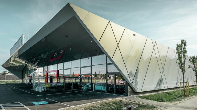 Der Eingangsbereich des EUROSPAR Supermarkts kombiniert großzügige Glasflächen mit einer markanten PREFA Aluminiumfassade. Die glänzende Oberfläche der Sonderfarbe Silver Gold unterstreicht die hochwertige, zukunftsorientierte Architektur der Supermarktfiliale. Die Aluminium Verbundplatten schillern in der Sonne je nach Blickwinkel in unterschiedlichen Farben.