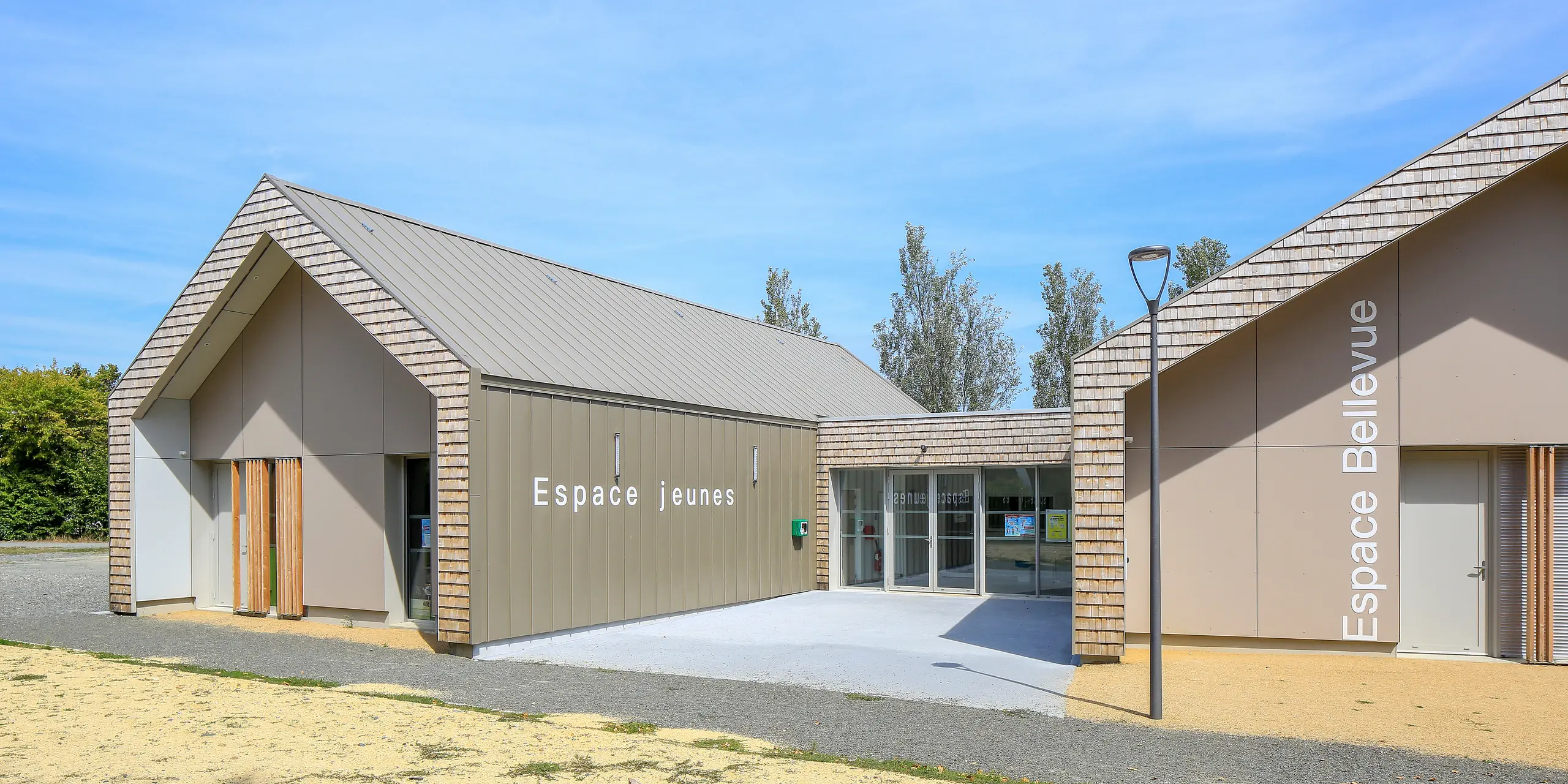 Salles Espace Bellevue à Mûrs-Erigné, France, avec les panneaux 'Espace Jeunes' et 'Espace Bellevue' devant la salle d'événements. Le système de toiture et de façade PREFALZ en bronze décore de grandes parties du centre de jeunesse moderne. Le bardage à joint debout en aluminium résistant aux intempéries de PREFA offre une solution durable et moderne pour les projets de construction. La combinaison du revêtement en aluminium et de l'aspect bois naturel s'intègre harmonieusement dans l'environnement.