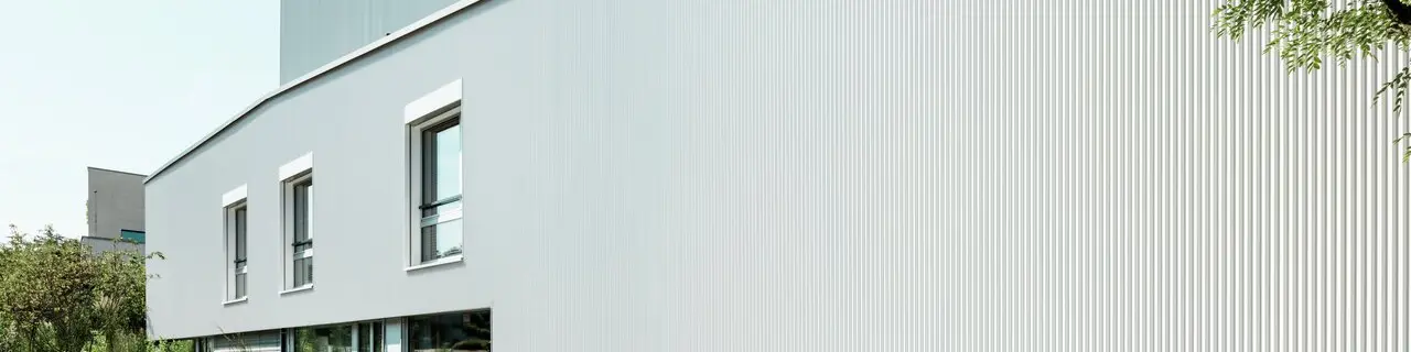 Vista moderna dell'edificio con facciata in PREFA con profilo a zeta in patina grigio. La struttura verticale del rivestimento in alluminio crea un vivace gioco di luci e ombre. Davanti all'edificio sono visibili finestre su due livelli, un'area verde e uno scooter bianco. Il cielo è limpido e azzurro.