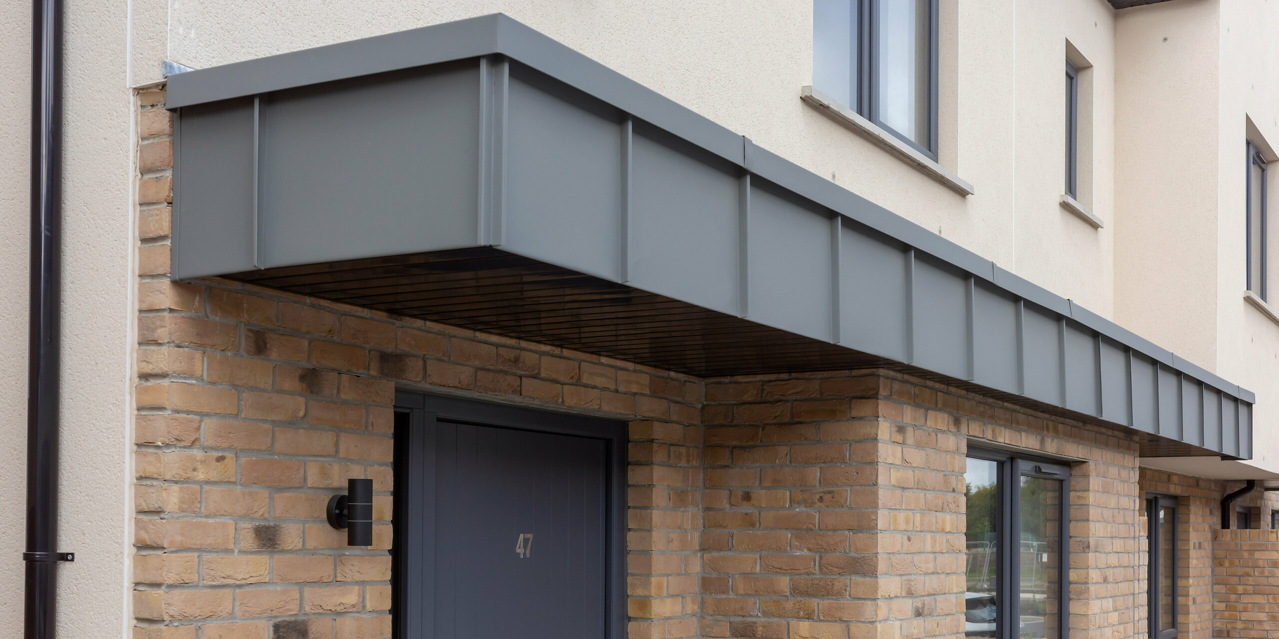 Le gros plan révèle l'entrée d'une maison mitoyenne à Churchfields, dont le porche d'entrée remarquable est revêtu de PREFALZ en coloris P.10 gris souris. Les lignes épurées et droites des surfaces en aluminium contrastent avec la chaleur de la brique et confèrent à l'entrée une profondeur architecturale de grande qualité. Résistante aux intempéries et inaltérable, la surface PREFA assure une protection durable et minimise l'entretien, ce qui est idéal pour les grands ensembles résidentiels. L'alliance du style métallique moderne et de la brique traditionnelle contribue à l'élégance générale de la scène.