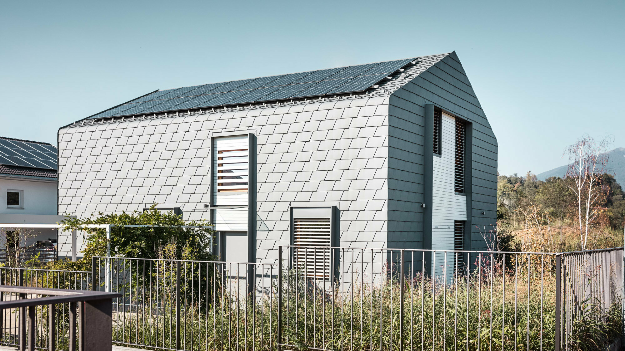 Vue latérale de la « Casa Longaretti », avec espace vert et clôture. La jonction arrondie entre toit et façade est bien visible ainsi que la façade en bardeaux gris souris.