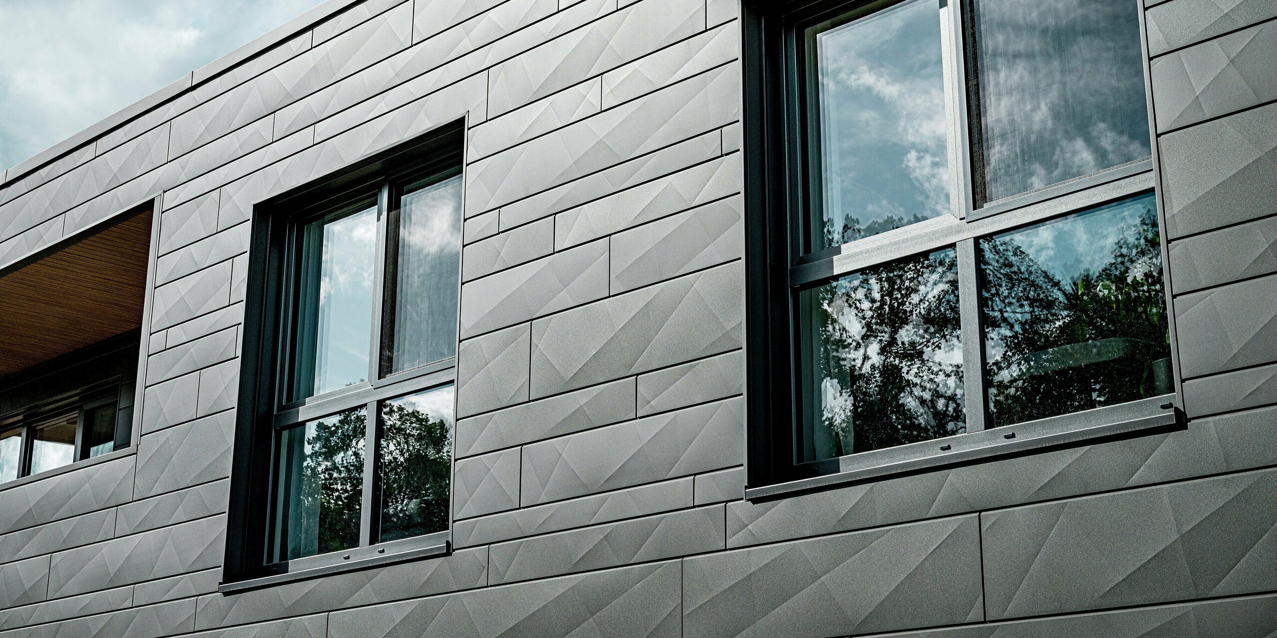 Nahaufnahme der PREFA Siding.X Aluminiumfassade in P.10 Dunkelgrau an einem modernen Einfamilienhaus in Heidenheim. Die unterschiedlich breiten Paneele mit markanten Kantungen erzeugen ein lebendiges, geometrisches Lichtspiel. Zwei große Fenster spiegeln Himmel und Baumkronen und betonen die klare, reduzierte Architektur. Der präzise Abschluss der Paneele zeigt die hohe Verarbeitungsqualität und die langlebige, witterungsbeständige Aluminiumoberfläche, die das Gebäude dauerhaft schützt und dessen moderne Optik unterstreicht.