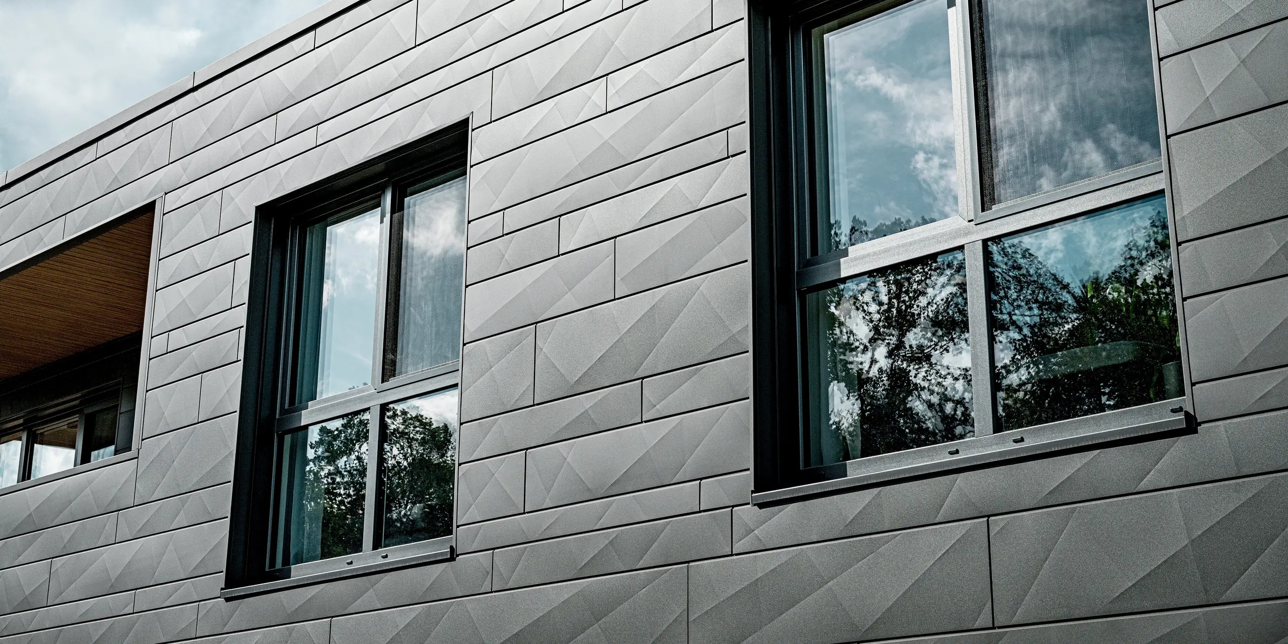 Gros plan sur la façade en aluminium Siding.X PREFA, coloris P.10 gris sombre, d'une maison individuelle moderne à Heidenheim. Les panneaux, de largeurs variables et aux arêtes marquées, créent un jeu de lumière géométrique et dynamique. Deux grandes fenêtres reflètent le ciel et la cime des arbres, soulignant l'architecture épurée et minimaliste. La finition précise des panneaux témoigne d'une fabrication de haute qualité et de la durabilité de la surface en aluminium, résistante aux intempéries, qui assure une protection durable au bâtiment et renforce son esthétique contemporaine.