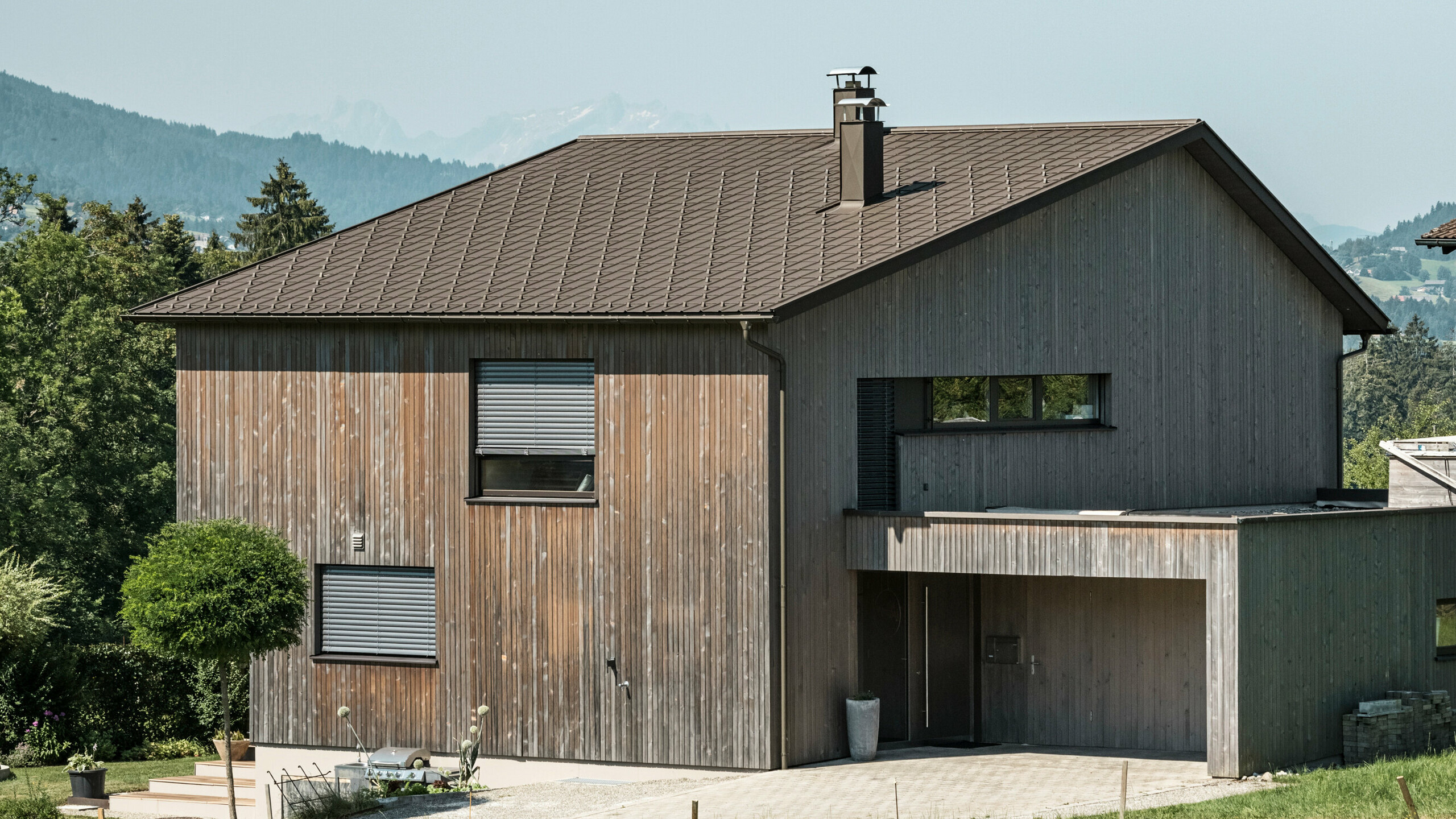 Vista laterale di una casa unifamiliare contemporanea a Krumbach con una combinazione di facciata in legno naturale e tetto in alluminio PREFA di lunga durata in P.10 marrone. L'armoniosa combinazione di colori mostra l'elegante integrazione di design moderno e materiali tradizionali, mentre le tegole PREFA assicurano un design del tetto estetico e funzionale.