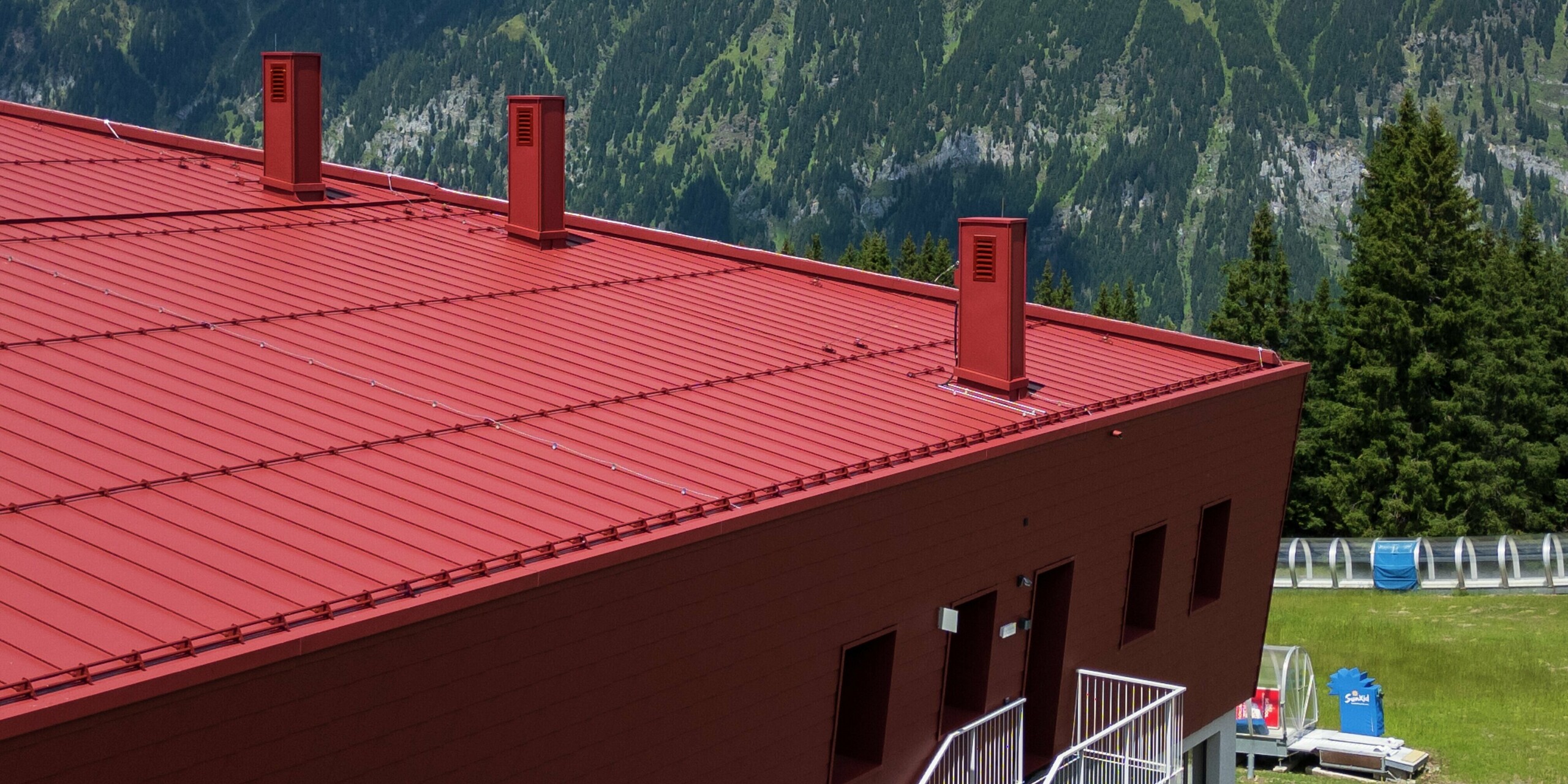 La toiture du Summit Mountain Club arbore la membrane à joints debout PREFALZ P.10 rouge oxyde, qui se fond harmonieusement dans le paysage alpin. Le système de ventilation performant et les lignes épurées de la toiture garantissent non seulement une esthétique soignée, mais aussi une fonctionnalité optimale. Ce matériau durable protège le bâtiment des intempéries et s'accorde parfaitement avec la façade remarquable qui se détache sur son environnement montagneux.