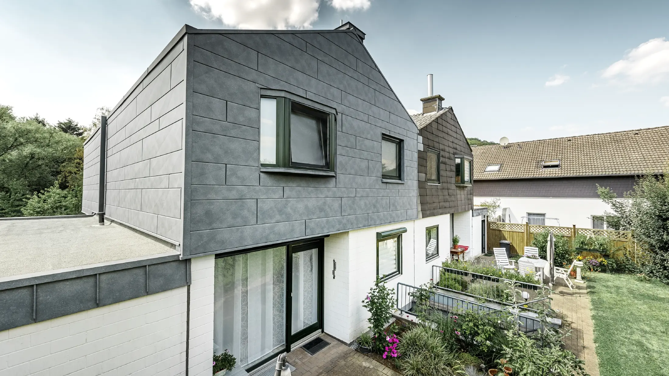 La parte superiore dell’edificio della metà di una casa bifamiliare è stata ristrutturata con l’elemento per facciata doga.X PREFA in P.10 grigio pietra. Accanto, a confronto, la metà della casa prima della ristrutturazione.