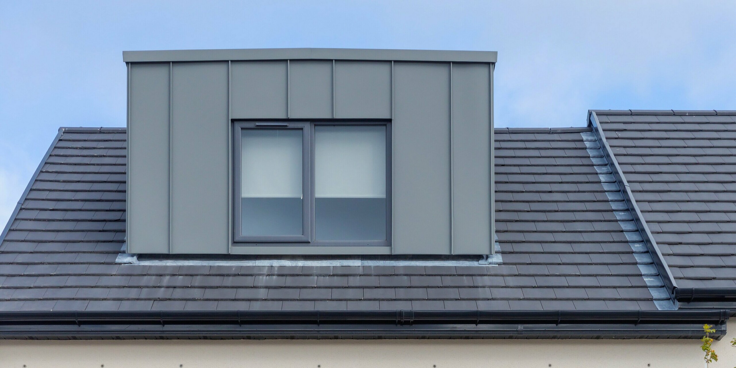 L'image présente une maison mitoyenne moderne à Churchfields, dont la lucarne est revêtue d'un bardage PREFALZ de haute qualité, coloris P.10 gris souris. La structure à joints debout épurés de la surface en aluminium crée un contraste saisissant au-dessus du toit de tuiles foncées et confère au bâtiment une allure élégante et contemporaine. Durable et résistant à la corrosion, le bardage PREFA garantit une esthétique harmonieuse et réduit considérablement l'entretien – un atout majeur pour les maisons de cette taille.
