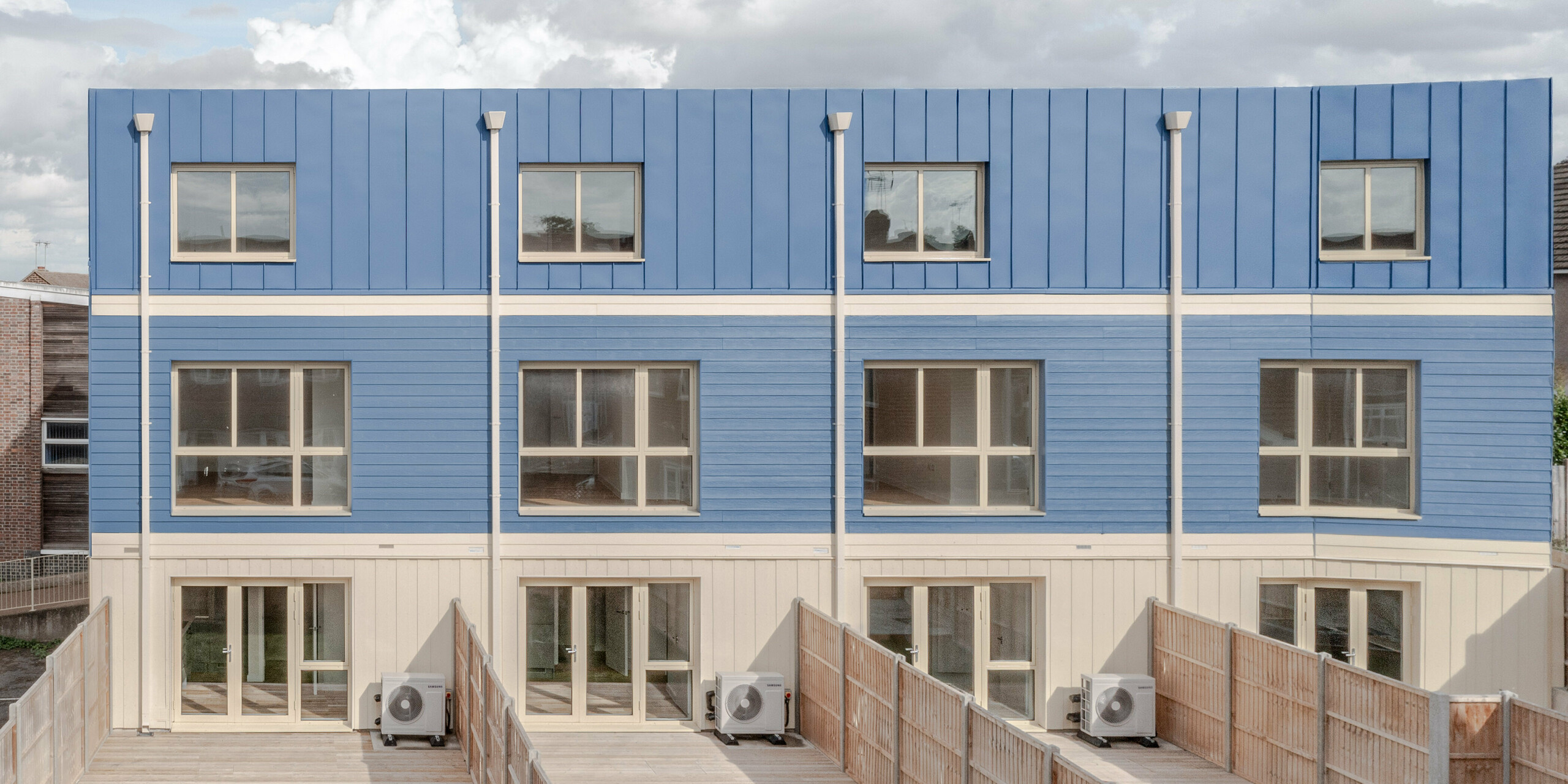 Die Gartenseite der Reihenhäuser in Goldsmith Mews zeigt die markante taubenblaue PREFA FALZONAL Fassaden- und Dachgestaltung, die den Gebäuden einen klaren, modernen Charakter verleiht. Die präzisen vertikalen Falze und die blauen Paneele erzeugen ein rhythmisches, lebendiges Fassadenbild. Die hellen Erdgeschossbereiche und großen Fenster öffnen die Architektur nach außen. Das hochwertige PREFA Aluminium bietet dauerhaften Witterungsschutz, hohe Farb- und Formstabilität und ermöglicht eine langlebige, ästhetische Gestaltung mit hoher Wiedererkennung.