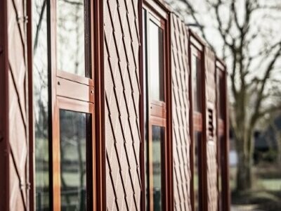 Detailaufnahme des Kindergartens, der seitlich fotografiert wurde. Die nebenliegenden Gebäude spiegeln sich in den Fenstern. Der Kindergarten Hennstedt wurde mit der Dachraute 29 × 29 in Oxydrot eingedeckt.